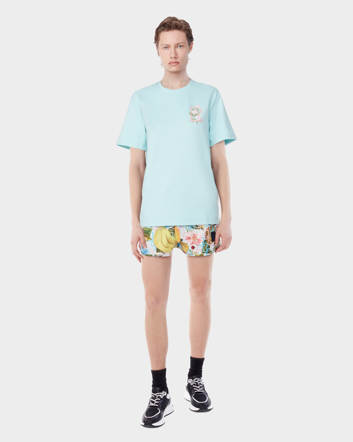 A person stands facing forward wearing the Rudsak DIAR - 9523003 M ICE BLUE T-shirt paired with colorful floral and banana print classic-cut shorts, black socks, and black sneakers against a plain white background.