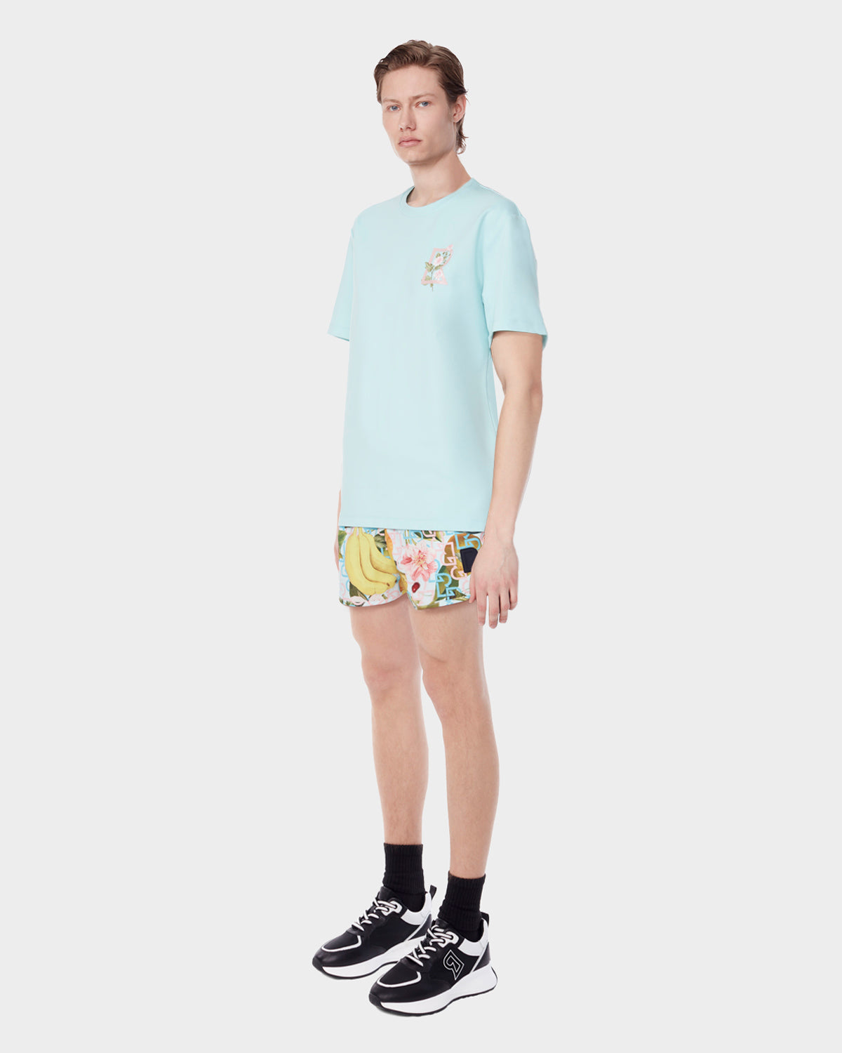 A person faces forward against a plain white background, wearing the Rudsak DIAR - 9523003 M ICE BLUE T-shirt with a small chest graphic, colorful tropical shorts, black socks, and black-and-white sneakers.