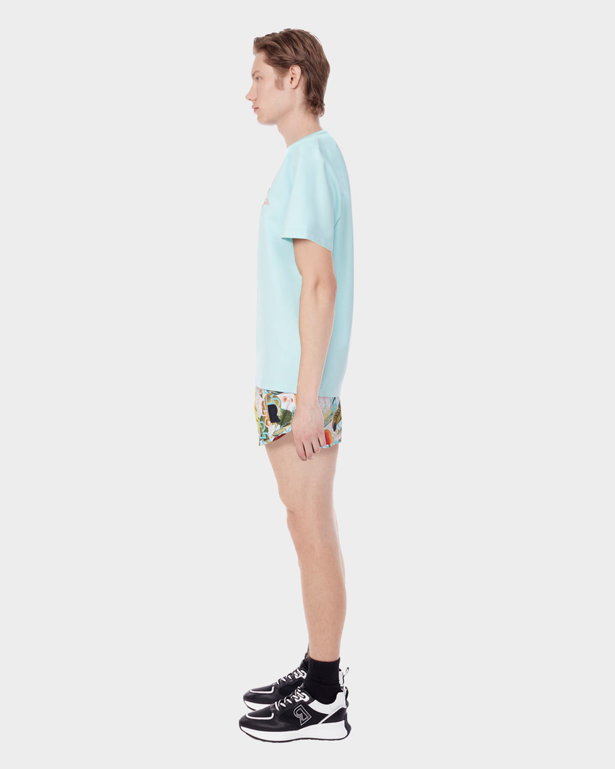 A person stands in profile against a plain white background, wearing the Rudsak DIAR - 9523003 M ICE BLUE t-shirt with a classic cut, colorful patterned shorts, black socks, and black sneakers.