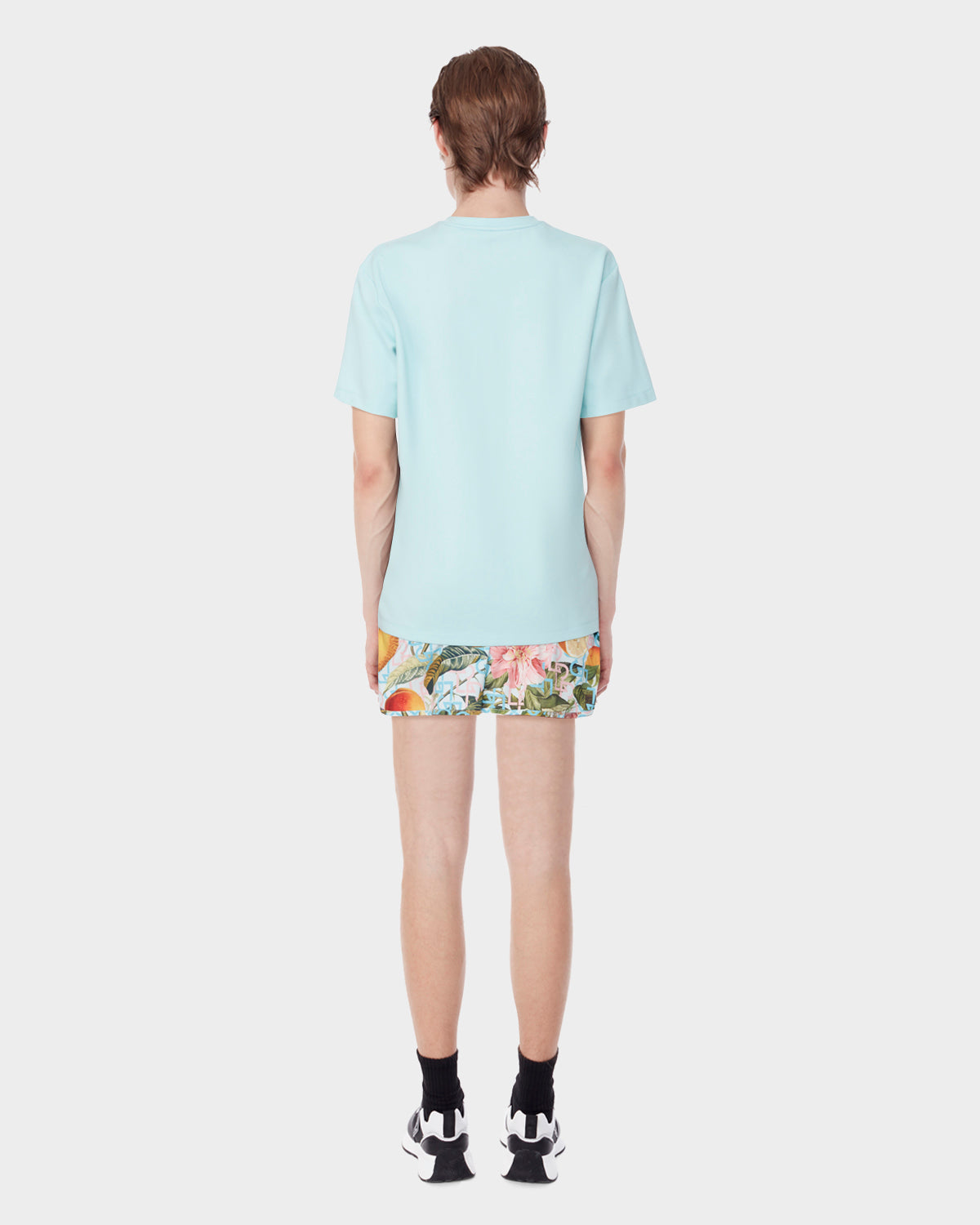 A person stands facing away from the camera, wearing the Rudsak DIAR - 9523003 M ICE BLUE short-sleeve shirt, colorful floral shorts, black socks, and black-and-white athletic shoes against a plain white background.