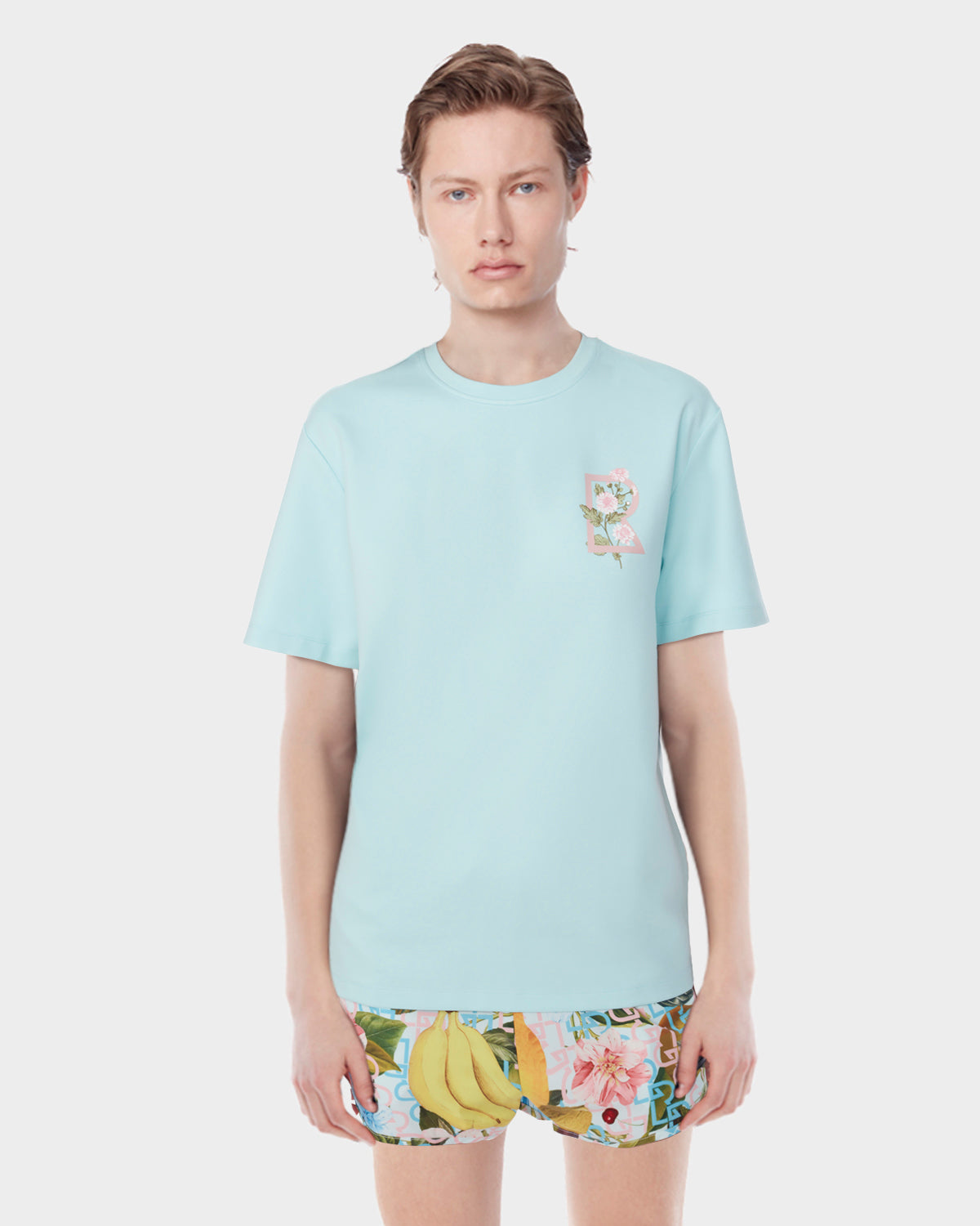 A person with short hair wears the Rudsak DIAR - 9523003 M ICE BLUE T-shirt, a classic light blue tee with a small floral graphic on the left chest, paired with colorful banana and floral print shorts against a white background.