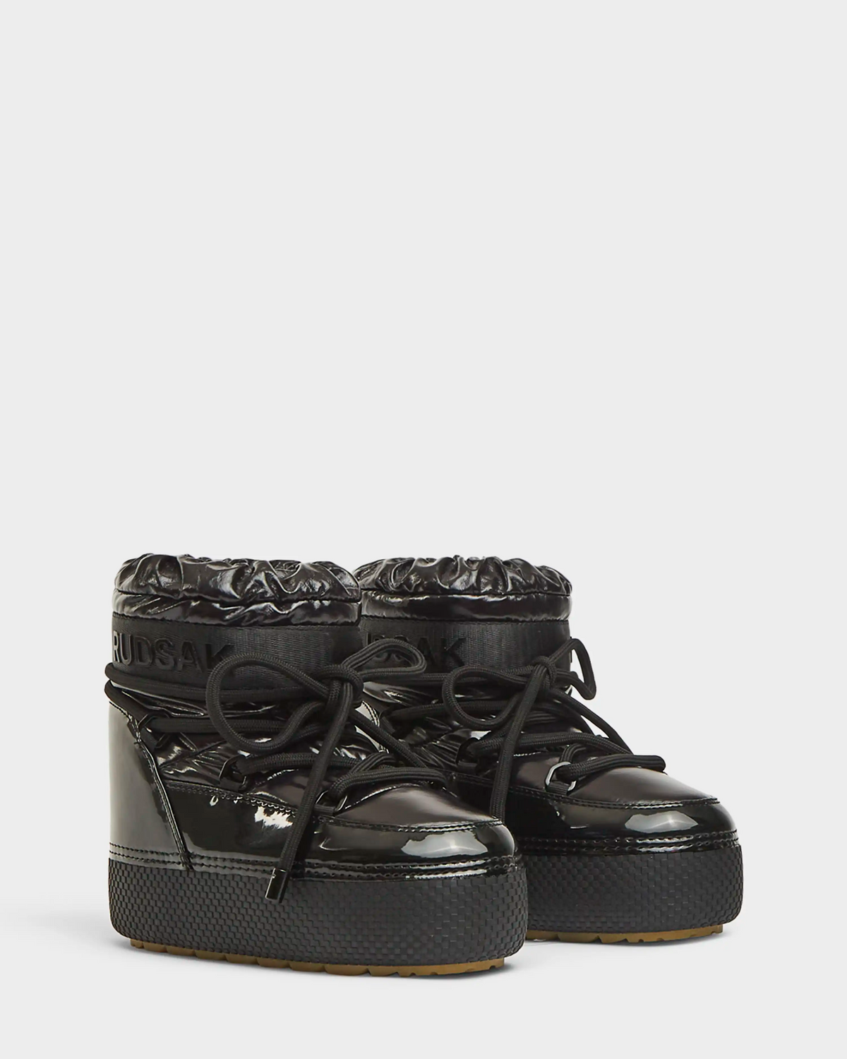 A pair of Rudsak MACE - 8223500 Black women's short moon boots with shiny, water-repellent quilted uppers, chunky laces, thick rubber soles, faux fur lining, and branded cuffs is displayed on a plain white background.