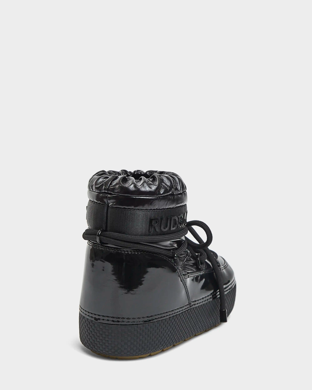 The MACE - 8223500 Black by Rudsak is a shiny black water-repellent women’s winter boot with a thick sole, visible laces, padded drawstring-style top, and “RUDSAK” embossed near the ankle. Shown from a rear side angle.
