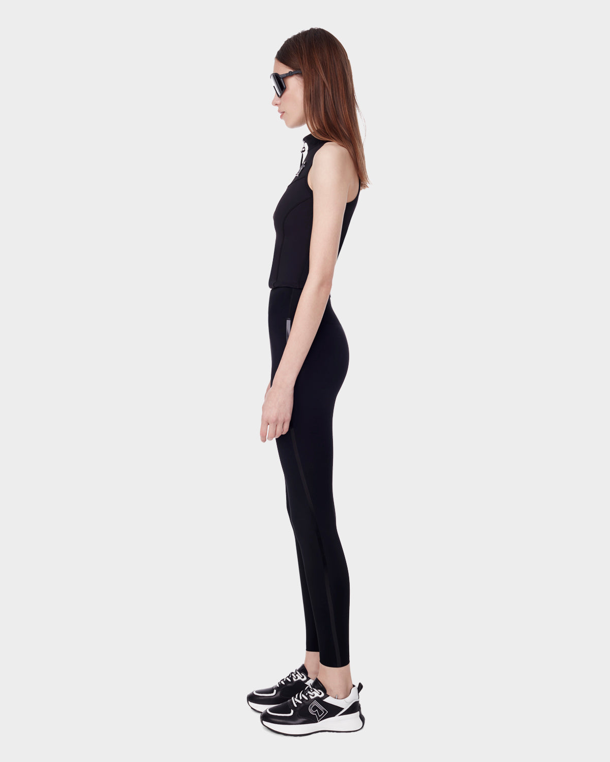 Woman in profile wears Rudsak’s KLEO F - 8523008 BLACK sleeveless top with high-rise technical stretch leggings, black-and-white sneakers, and sunglasses against a plain white background—ideal for an athleisure look.