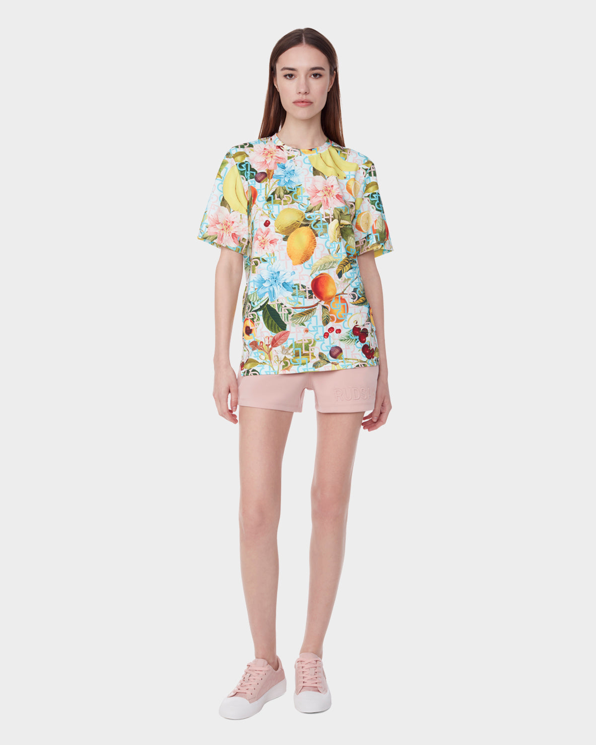 A woman faces forward against a white background wearing the Rudsak RAYA - 9523004 COCKTAIL RUDSAK F unisex T-shirt with a colorful fruit cocktail print, paired with light pink shorts and pale pink sneakers.