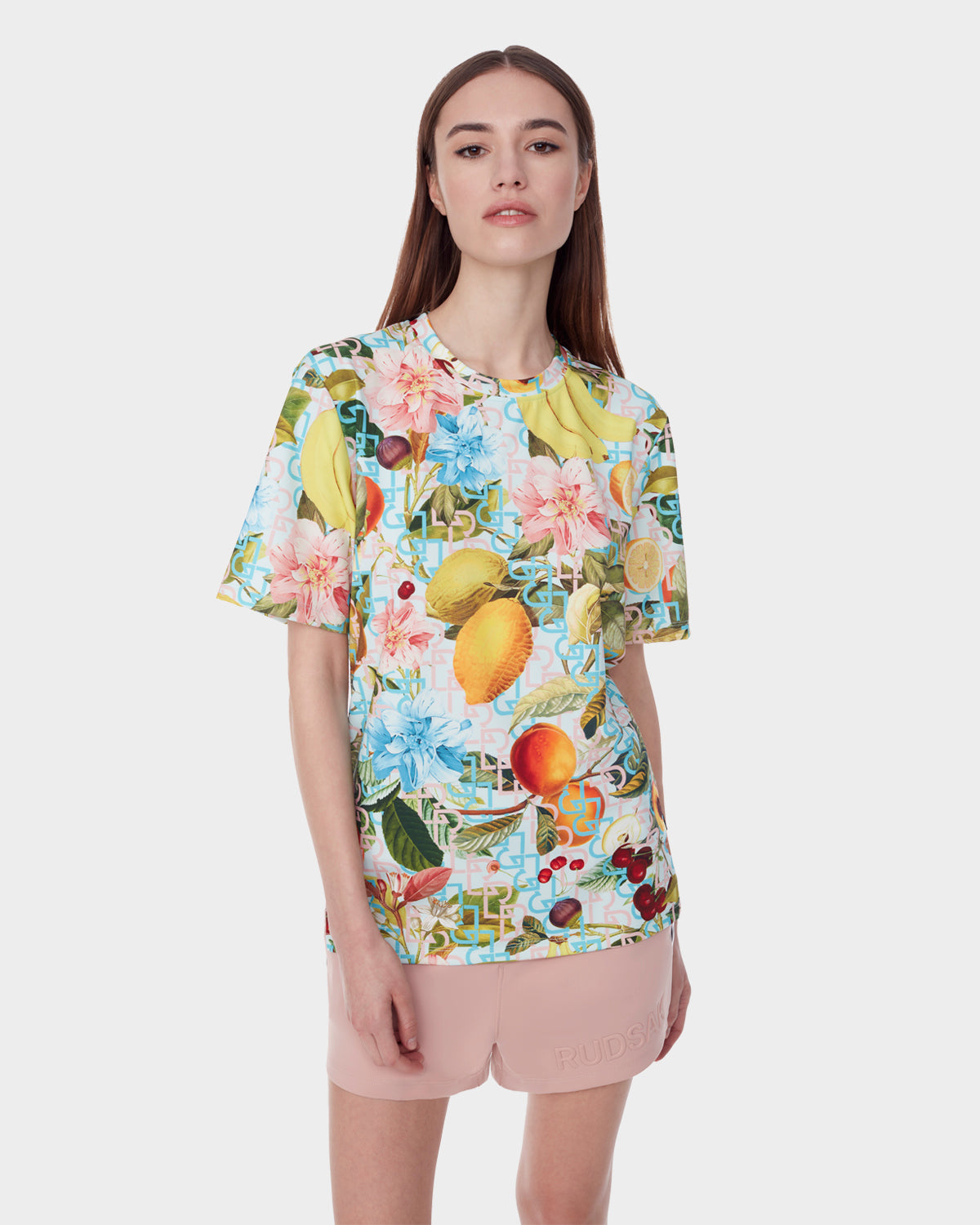 A woman with straight brown hair wears the RAYA - 9523004 COCKTAIL RUDSAK F T-shirt by Rudsak, which features a vibrant fruit cocktail and floral print, paired with pale pink shorts against a plain white background.