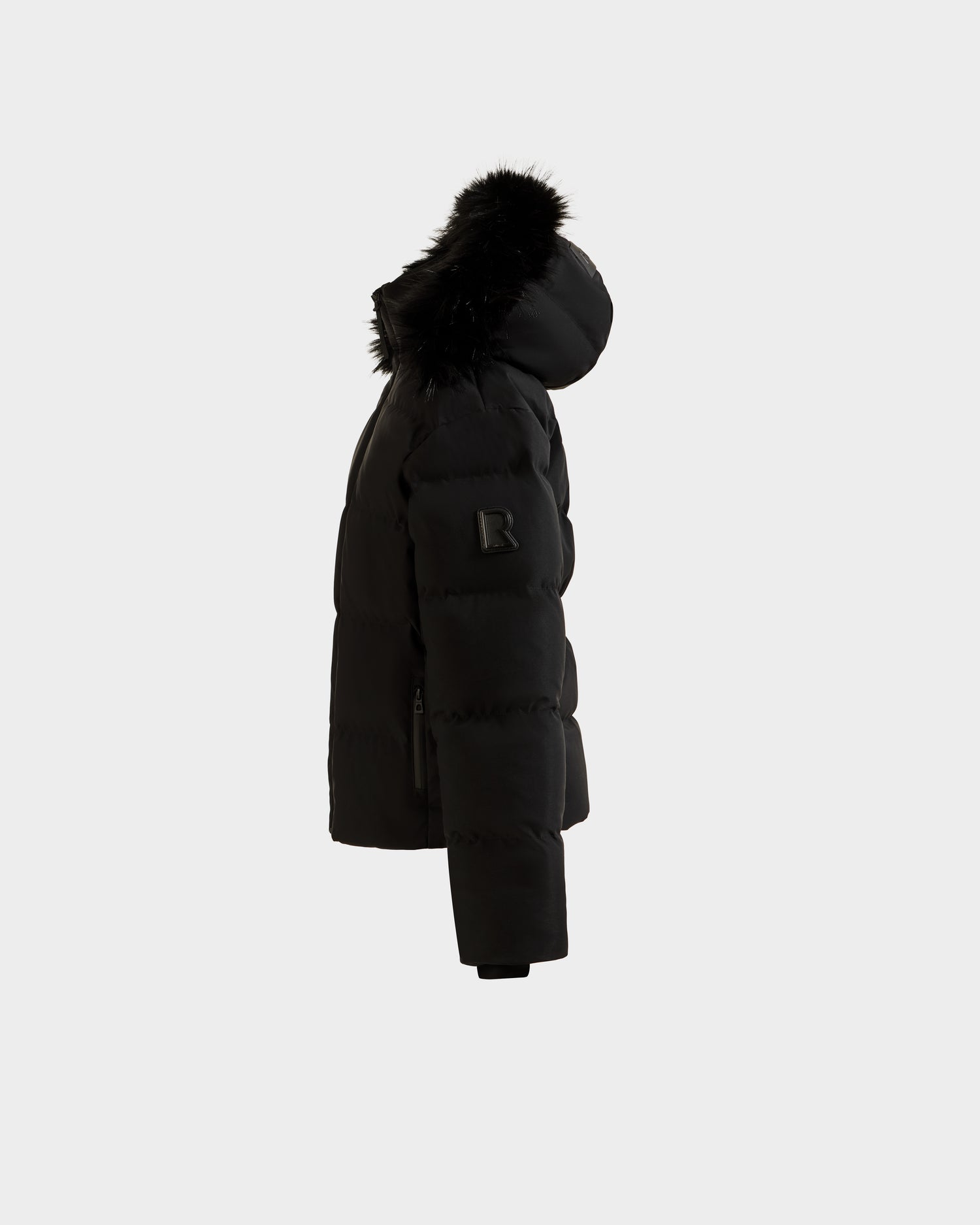 Side view of the Rudsak RAEGAN - 0125503 Black puffer jacket, featuring a hood with black faux fur trim, a sleeve zipper pocket, and stitched patch detail, shown against a plain light gray background.