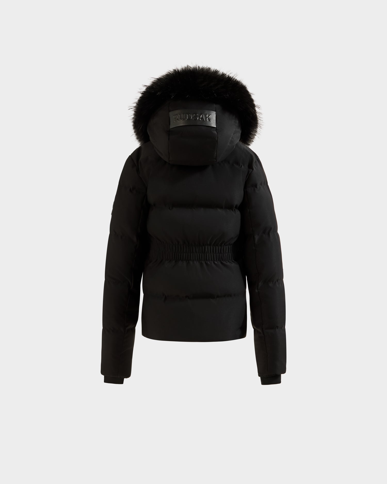 The RAEGAN - 0125503 Black by Rudsak is a black puffer jacket featuring a cinched waist and a hood trimmed with black fur, shown from the back against a plain white background. The jacket appears thick and warm.