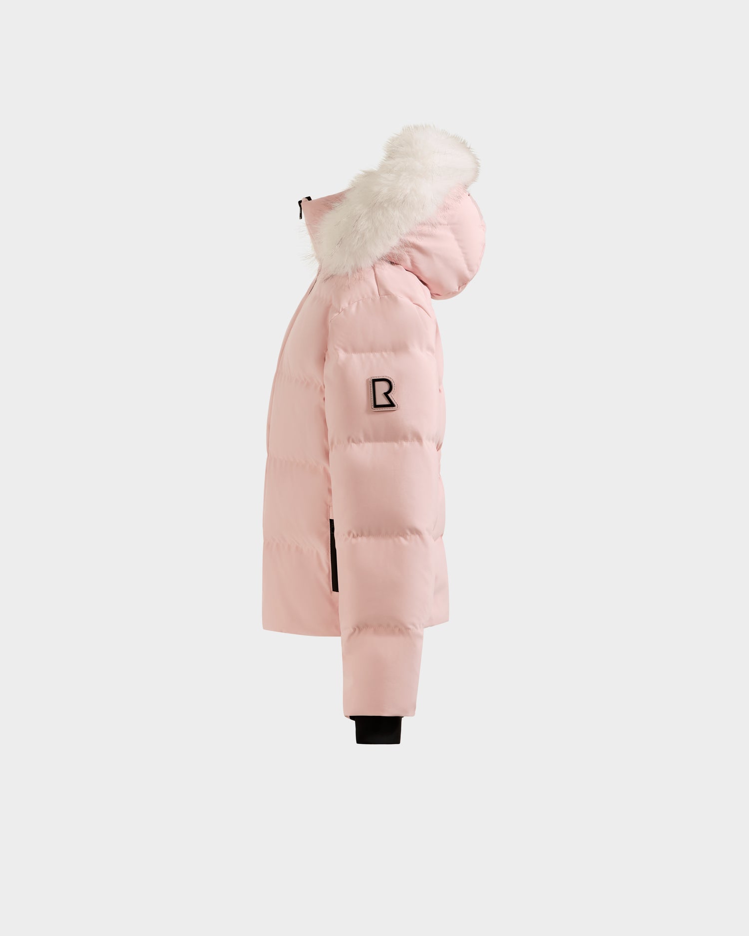 The Rudsak RAEGAN - 0125503 Pastel Pink puffer jacket features a hood with white faux fur trim, black zipper pockets, and a black B patch on the sleeve, shown from the side against a white background.