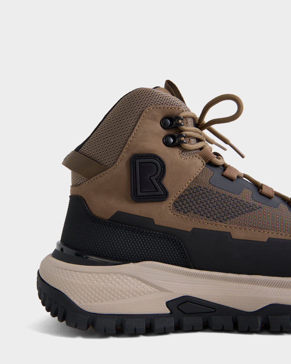 A close-up of the Rudsak REXON - 6226503 Cement winter-ready boot’s side in tan and black displays textured materials, sturdy laces, a rugged sole, and a prominent raised R logo. Only the rear and mid sections of this high-top sneaker are visible against a white background.