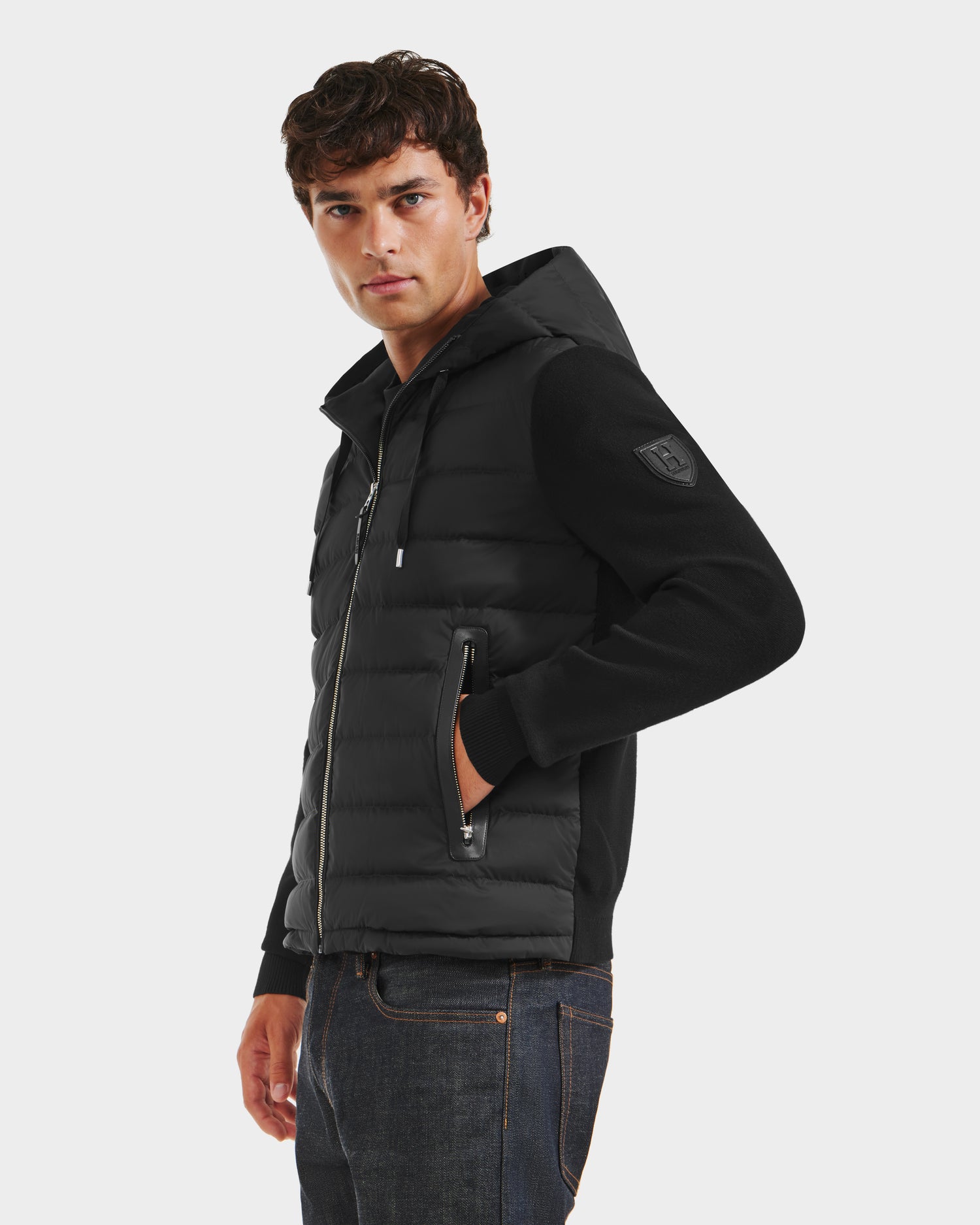 A young man wears the Rudsak RIVER HV - H125503 Black limited edition hybrid down jacket with zippered pockets and dark jeans, standing against a plain white background and looking slightly to the side.