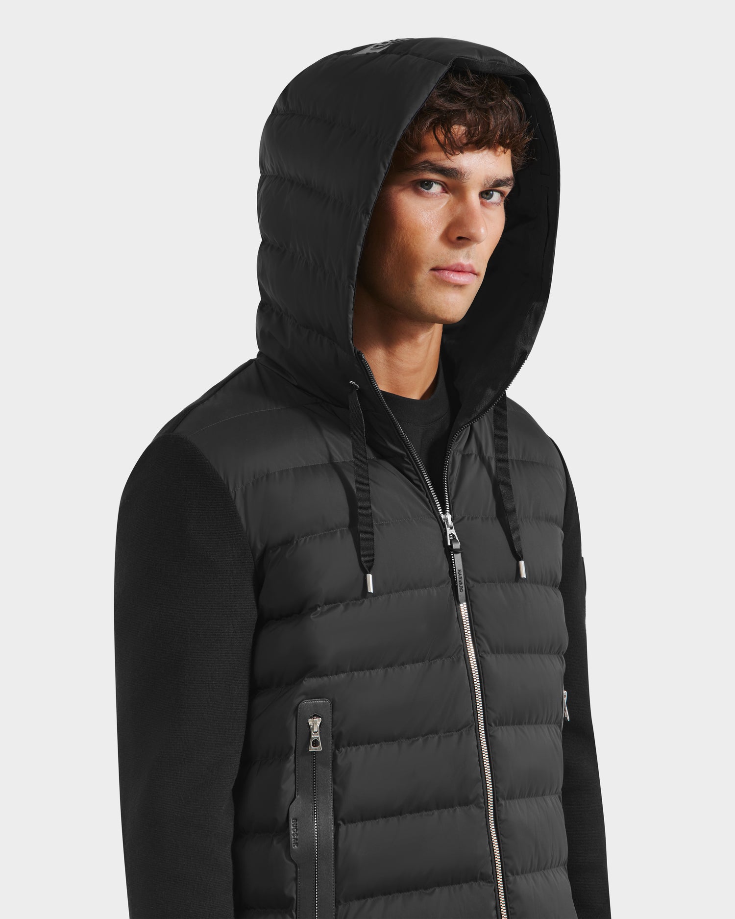 A young man with short brown hair wears the Rudsak RIVER HV - H125503 Black hybrid down jacket with a hood over a black shirt, standing against a plain light background and gazing slightly to the side.
