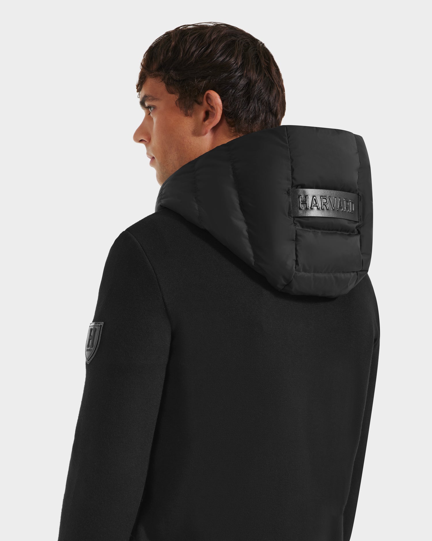 A man with short brown hair is seen from the back and side against a plain background, wearing the Rudsak RIVER HV - H125503 Black coat, a limited edition style with a padded hood and HARVII stitched in black, plus an upper arm patch.