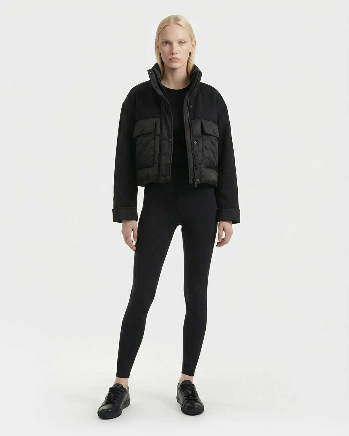 A person with long blonde hair models the Rudsak ROME - 8126028 Black cropped mixed-media jacket, styled with black leggings and sneakers against a plain white background.
