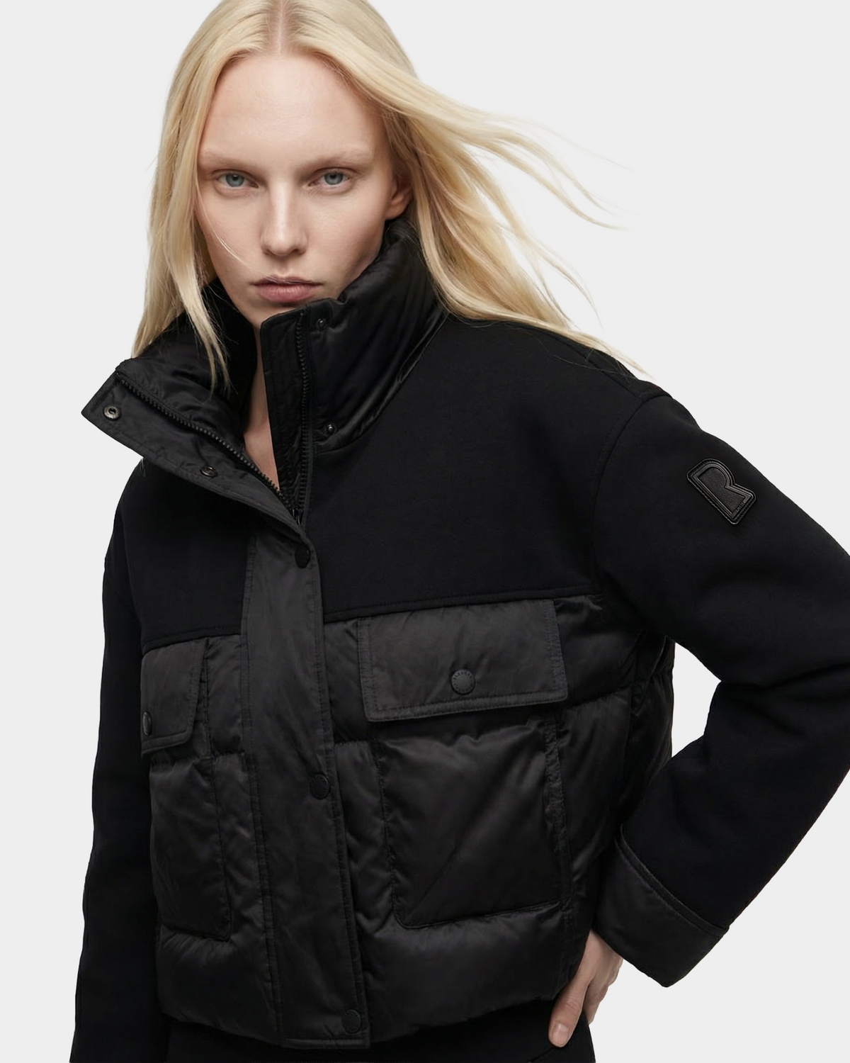 A woman with long blonde hair models the Rudsak ROME - 8126028 Black puffer jacket, featuring large front pockets and a B patch on the sleeve, posing with one hand on her hip against a plain background.