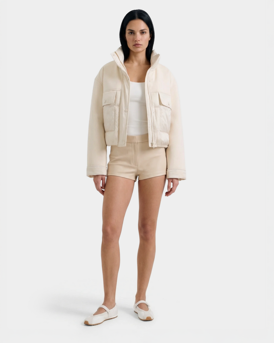 A woman faces forward against a plain white background, wearing the Rudsak ROME - 8126028 Iceland cream cropped spring jacket, a white tank top, beige shorts, and white strappy flat shoes.