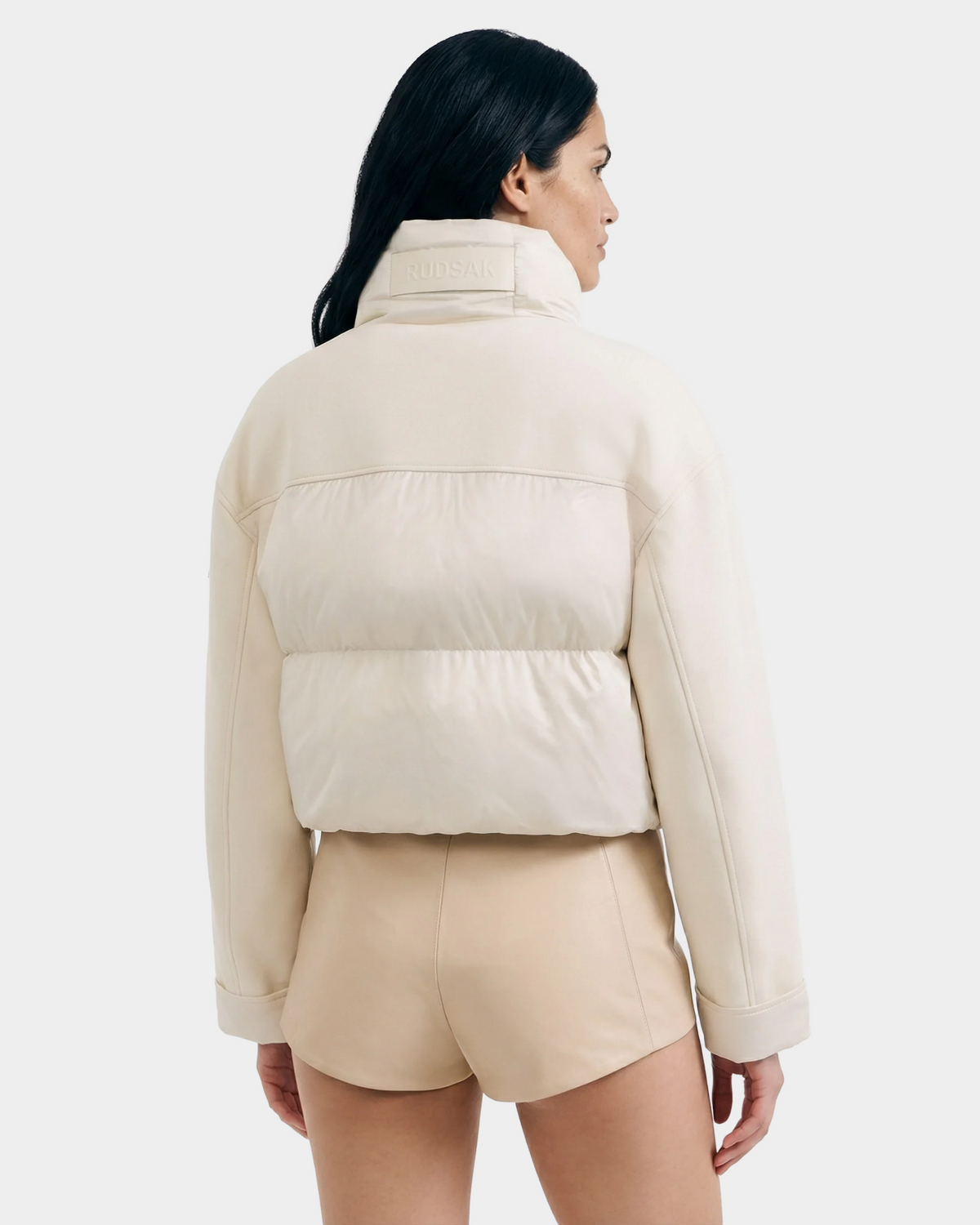 A woman with long dark hair wears the ROME - 8126028 Iceland light beige quilted down-filled jacket and matching shorts by Rudsak, standing with her back to the camera against a plain light background.