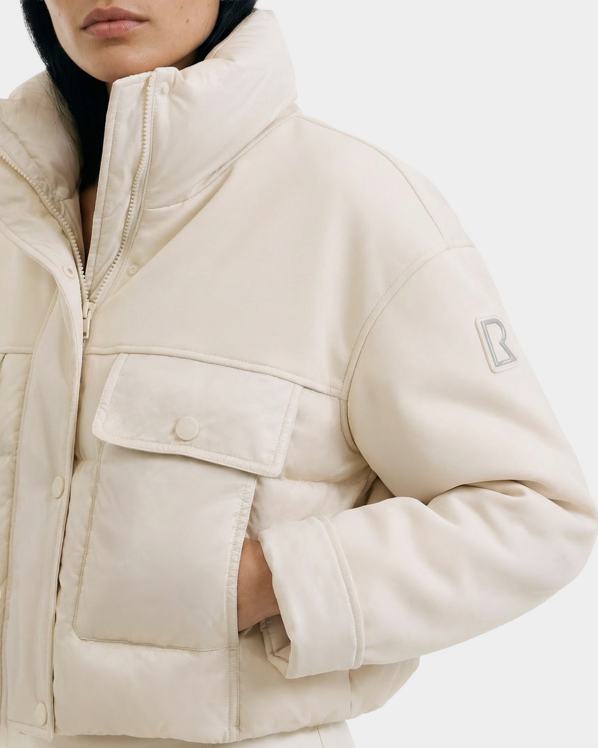 A person wears the Rudsak ROME - 8126028 Iceland light beige quilted down jacket with large front pockets, a high collar, and an R patch on the upper left sleeve. The left hand is tucked in a pocket. Background is plain white.