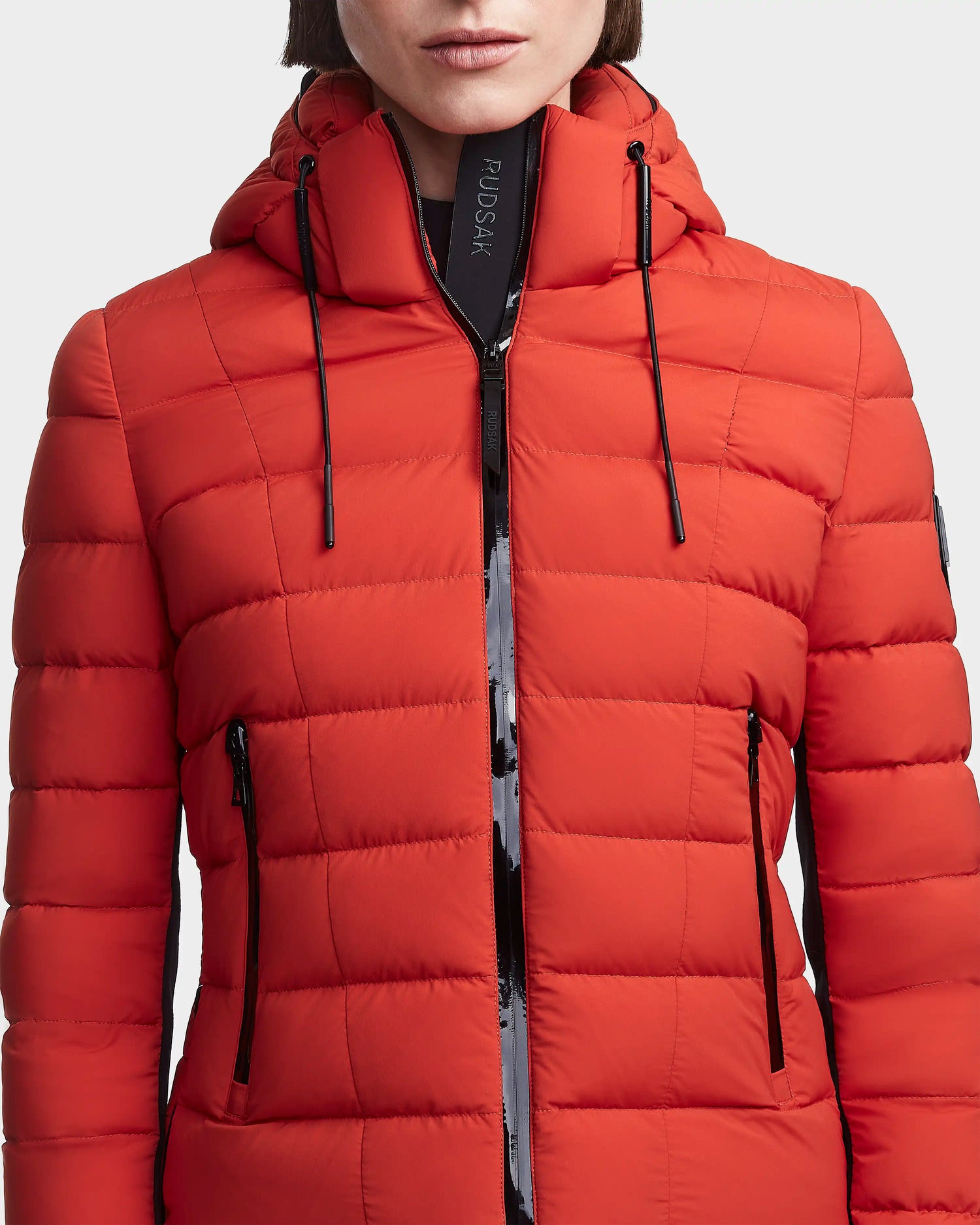 A woman wears the MELISSA - 8122551 Orange puffer jacket by Rudsak, featuring black zipper accents and a hood, shown from shoulders to waist against a plain light background.
