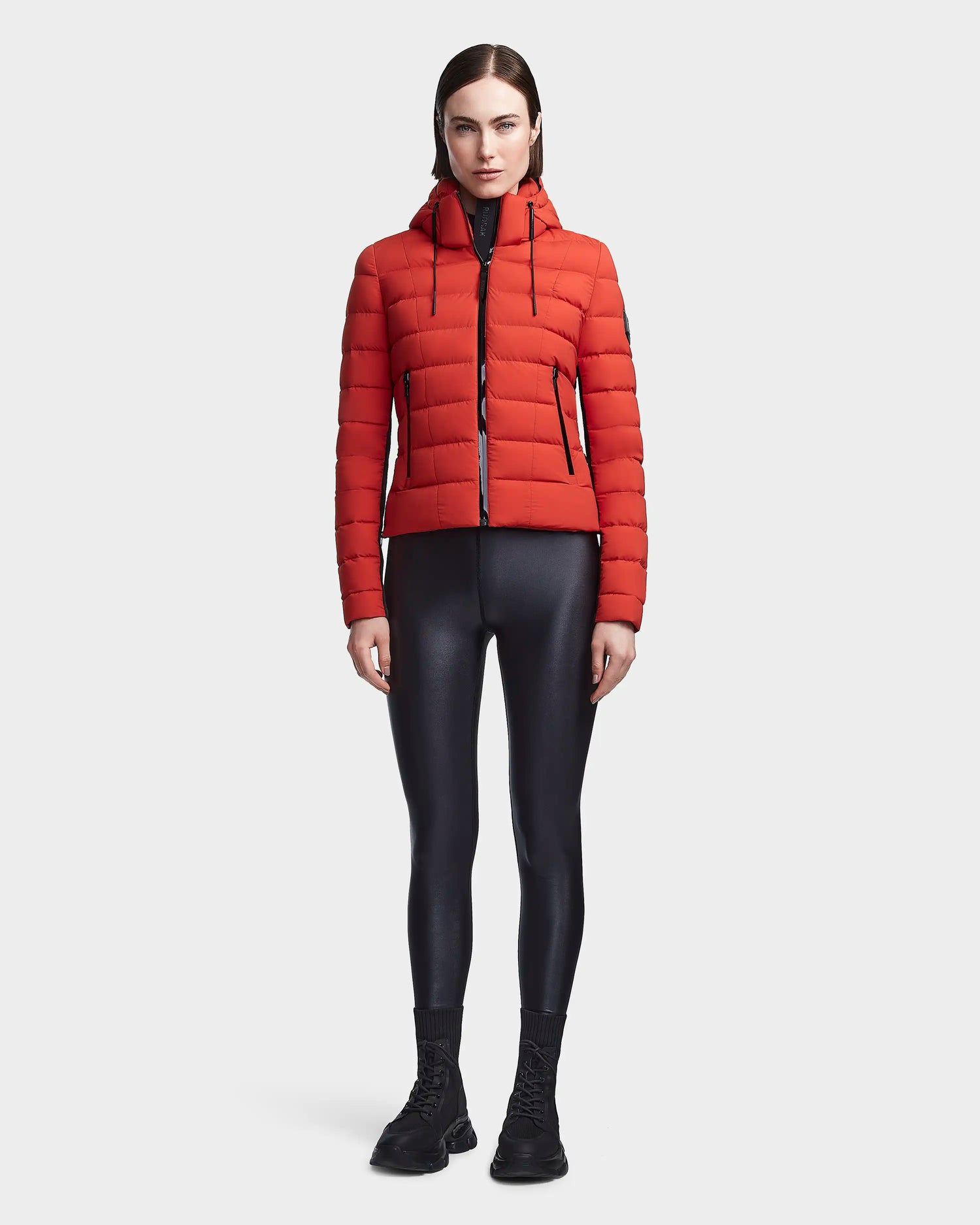 A woman stands facing forward wearing the Rudsak MELISSA - 8122551 Orange puffer jacket, paired with black shiny leggings, socks, and shoes against a plain white background.