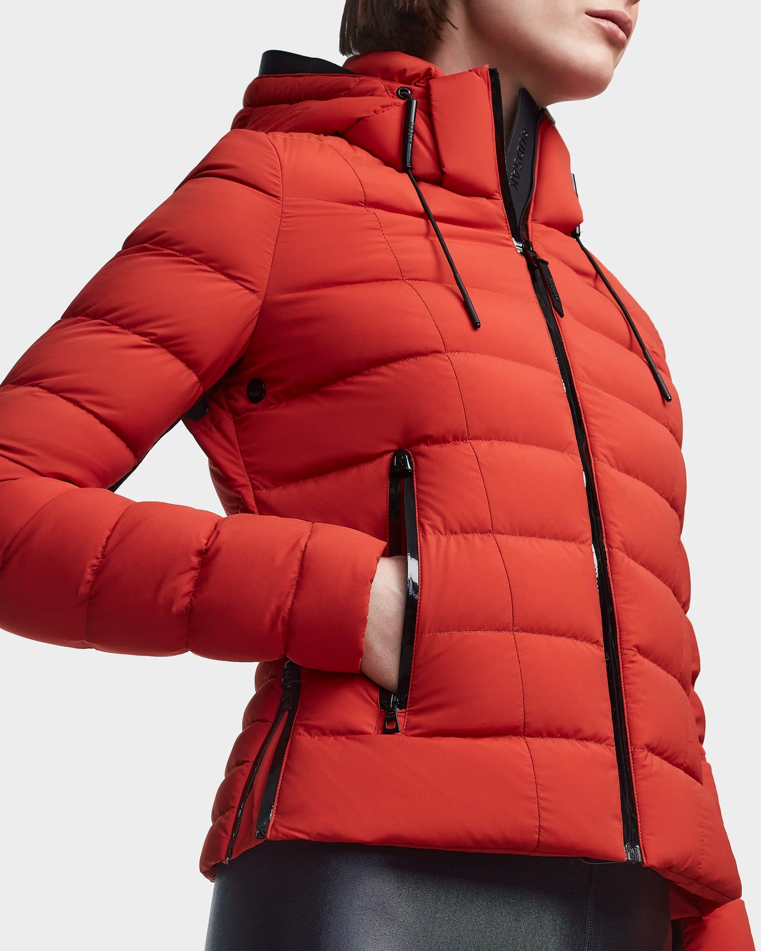 A person is wearing the Rudsak MELISSA - 8122551 Orange women's puffer jacket with black zippers and a hood, shown from shoulders to hips, one hand in the pocket. This stylish lightweight down jacket pops against a plain background.