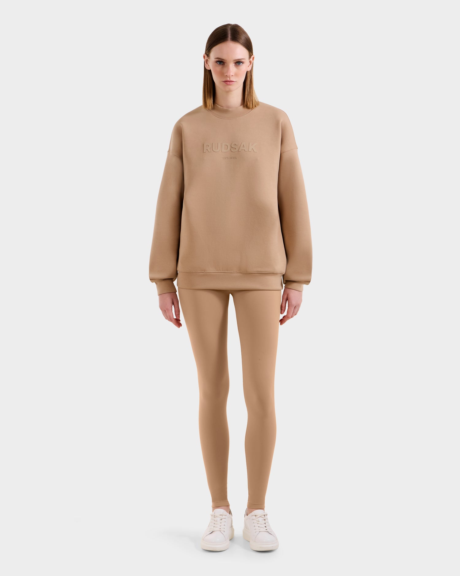 A woman faces forward in the BRYN - 9524007 Almond W oversized cotton crewneck sweatshirt by Rudsak, paired with matching beige leggings and white sneakers, set against a plain white background.