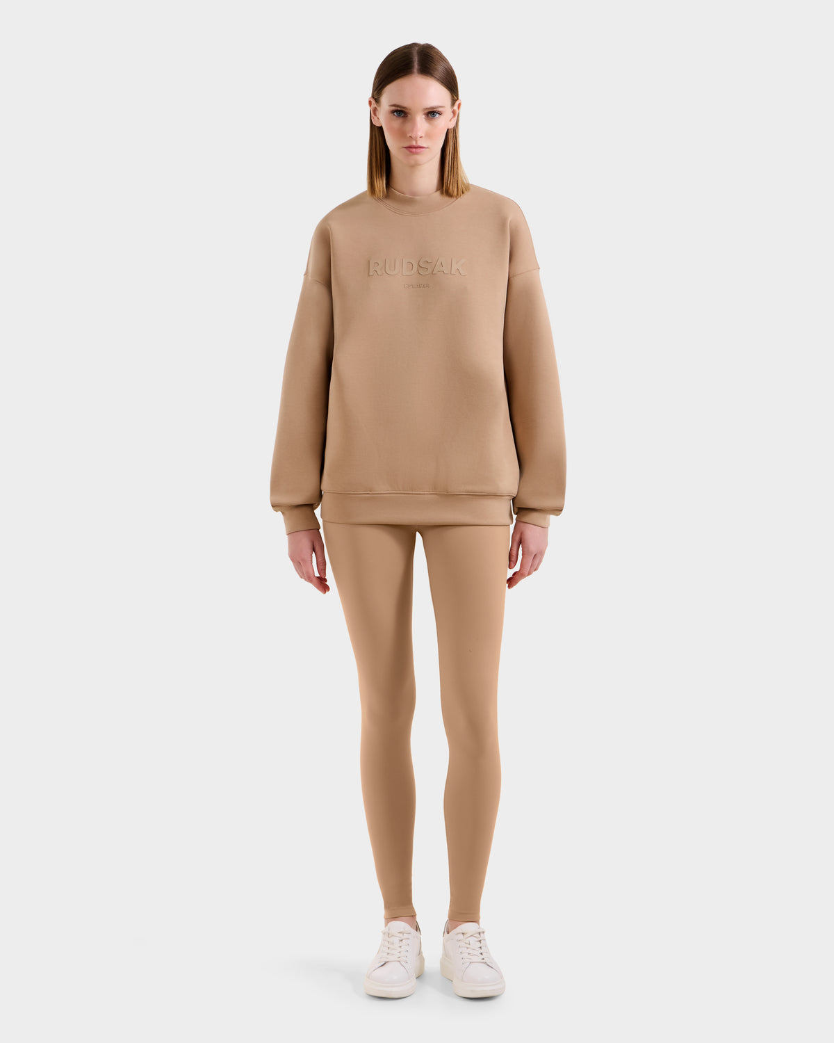 A woman faces forward in the BRYN - 9524007 Almond W oversized cotton crewneck sweatshirt by Rudsak, paired with matching beige leggings and white sneakers, set against a plain white background.
