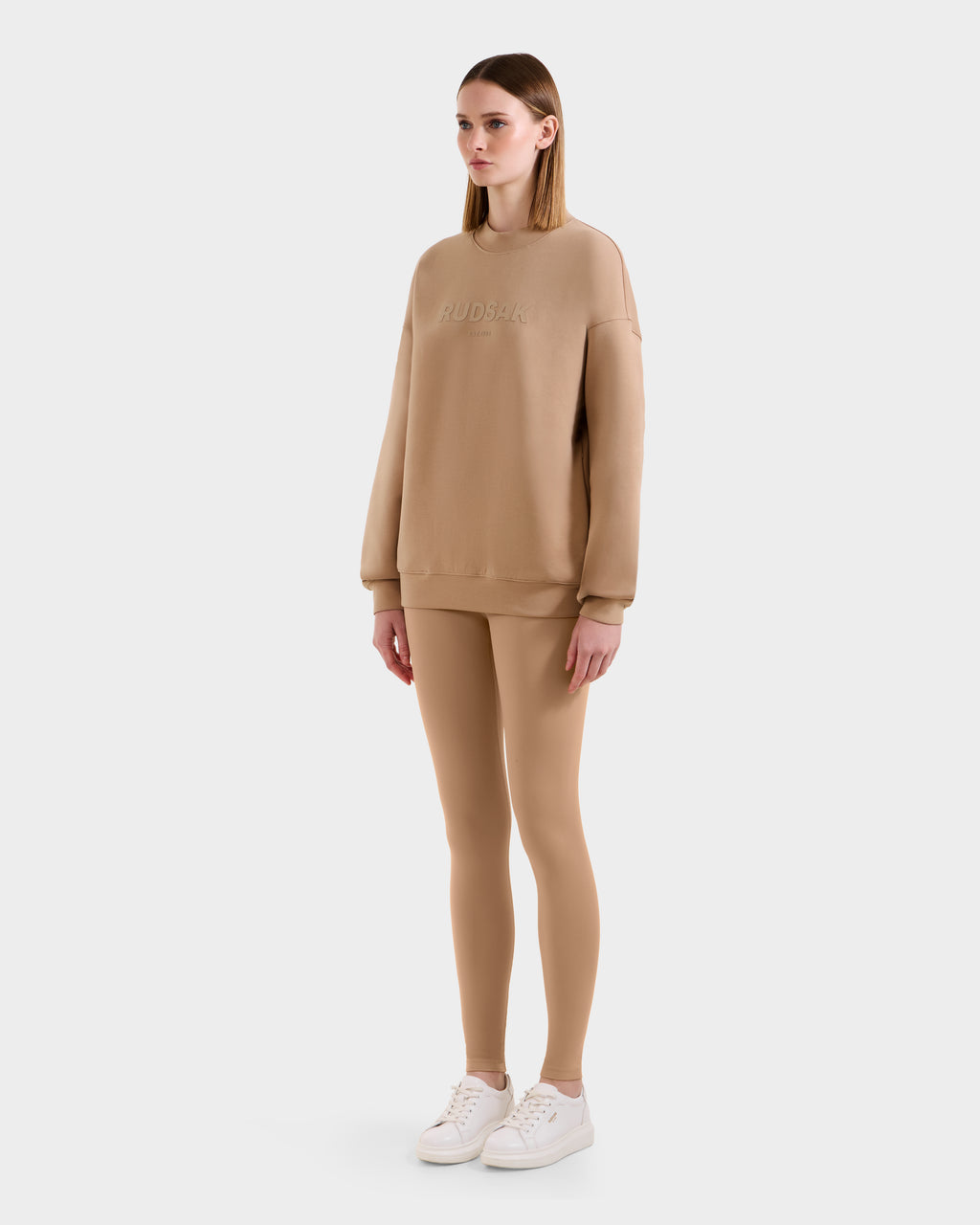 A woman faces forward in a Rudsak BRYN - 9524007 Almond W tan cotton crewneck sweatshirt and matching leggings with white sneakers. Her straight, loose hair and the plain light gray background highlight this ideal everyday look.