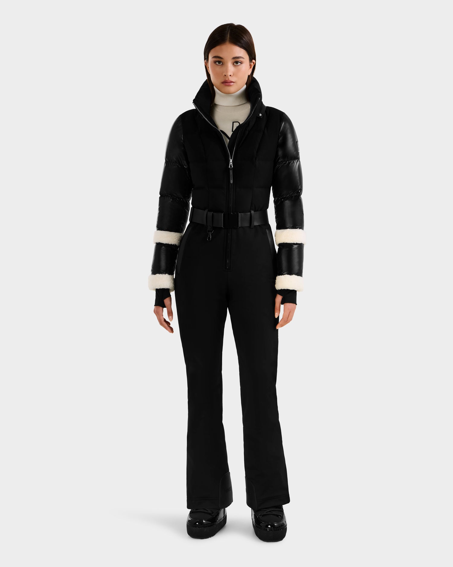 A woman stands facing forward in the Rudsak CARMEN - 8125708 Black fitted ski suit and puffer jacket with white fur sleeve trim, a white turtleneck, black gloves, and black boots against a plain light background.