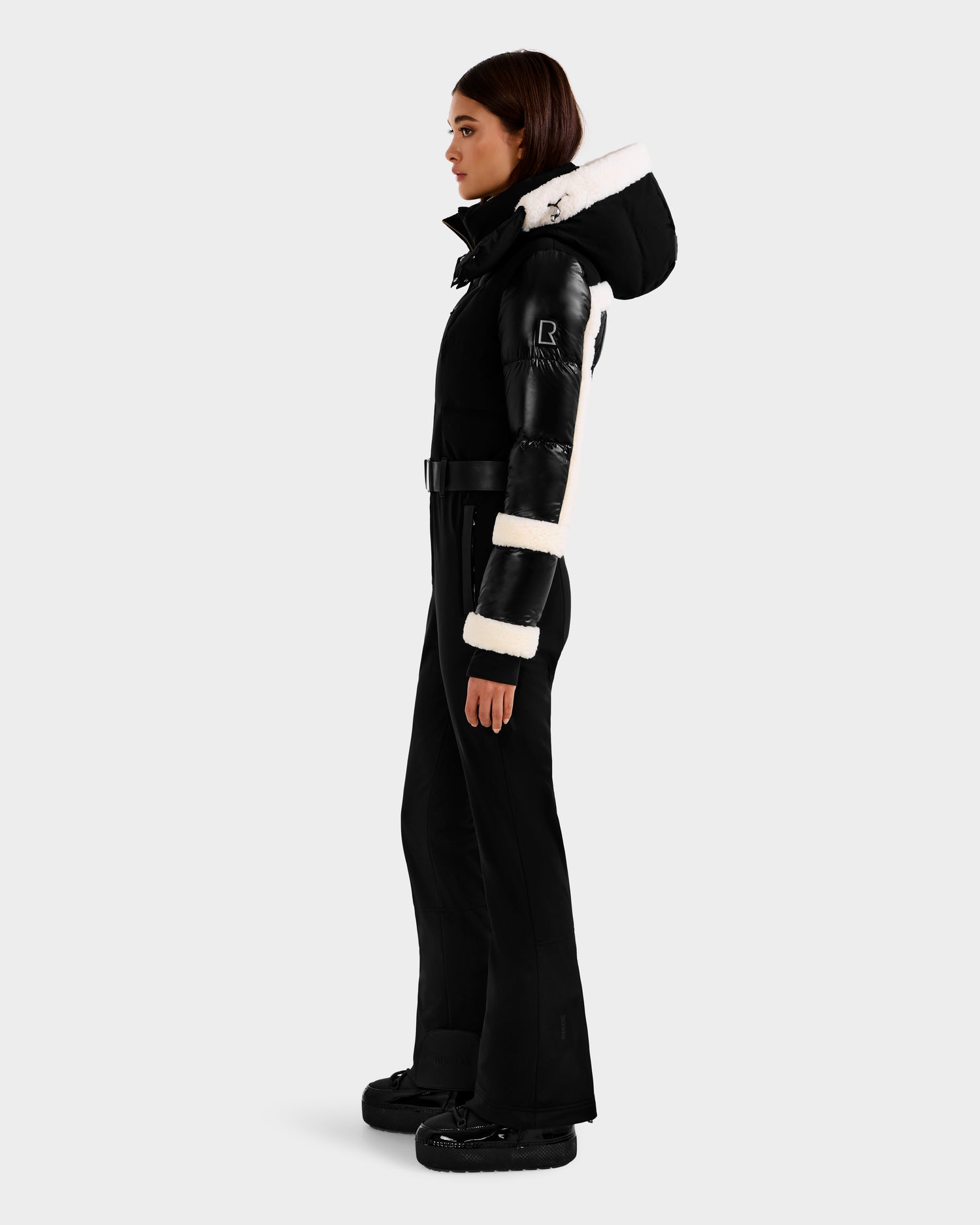 A woman in profile wears the Rudsak CARMEN - 8125708 Black fitted ski suit with white fur-trimmed hood and sleeves, a belt, and matching black boots, standing against a plain light gray background.