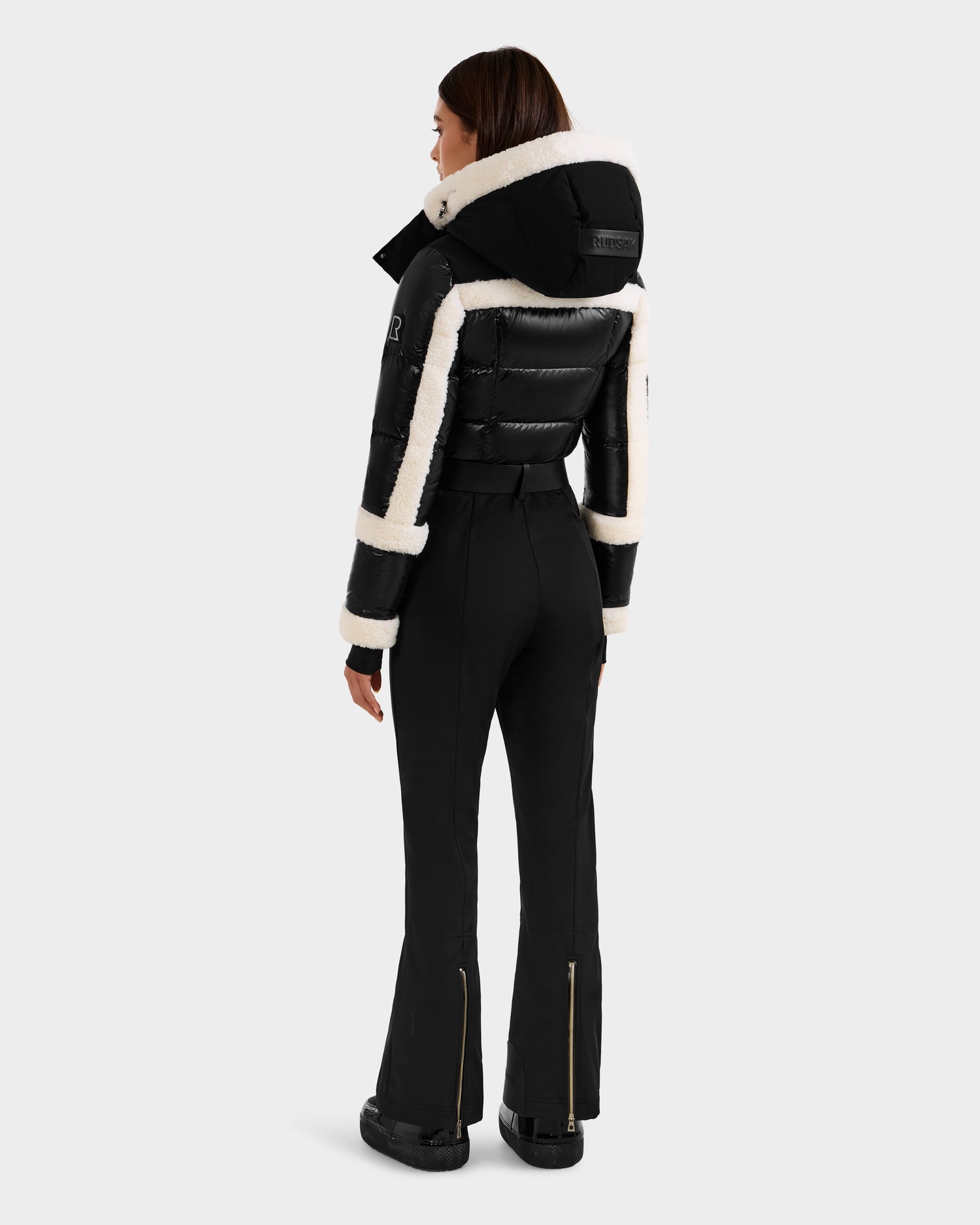 A woman faces away in the Rudsak CARMEN - 8125708 Black ski suit: a fitted black puffer jacket with white trim, matching high-waisted pants with gold ankle zippers, and black boots, set against a plain light background.