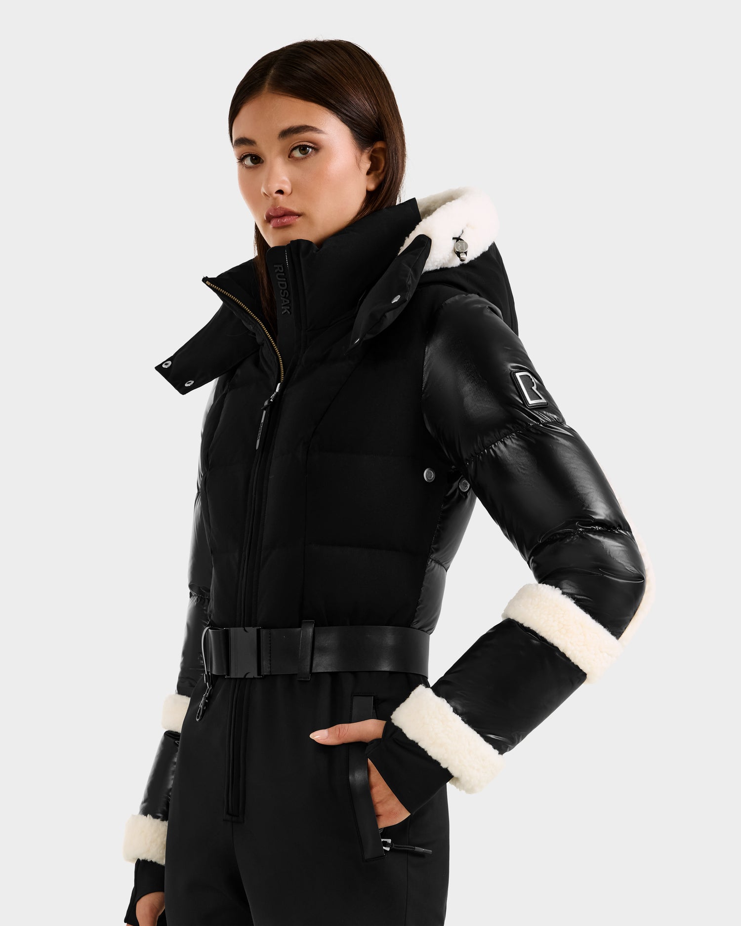 A woman models the Rudsak CARMEN - 8125708 Black puffer jacket with a belt, white fleece-trimmed hood and cuffs, and RECCO rescue technology, standing against a plain light gray background.