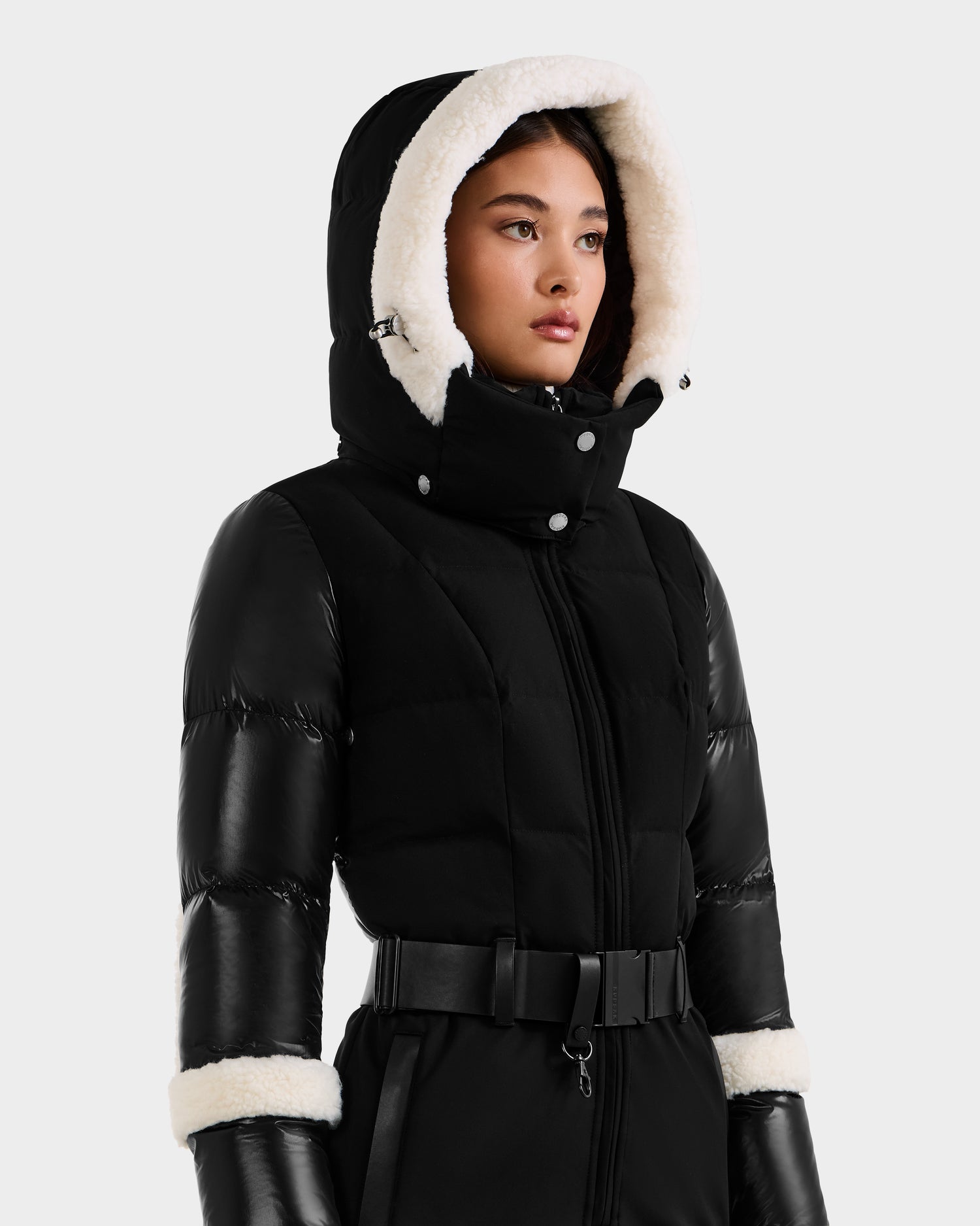 A woman wears the Rudsak CARMEN - 8125708 Black ski suit with a belted waist, white faux fur-trimmed hood and cuffs, and black gloves, standing against a plain light background.