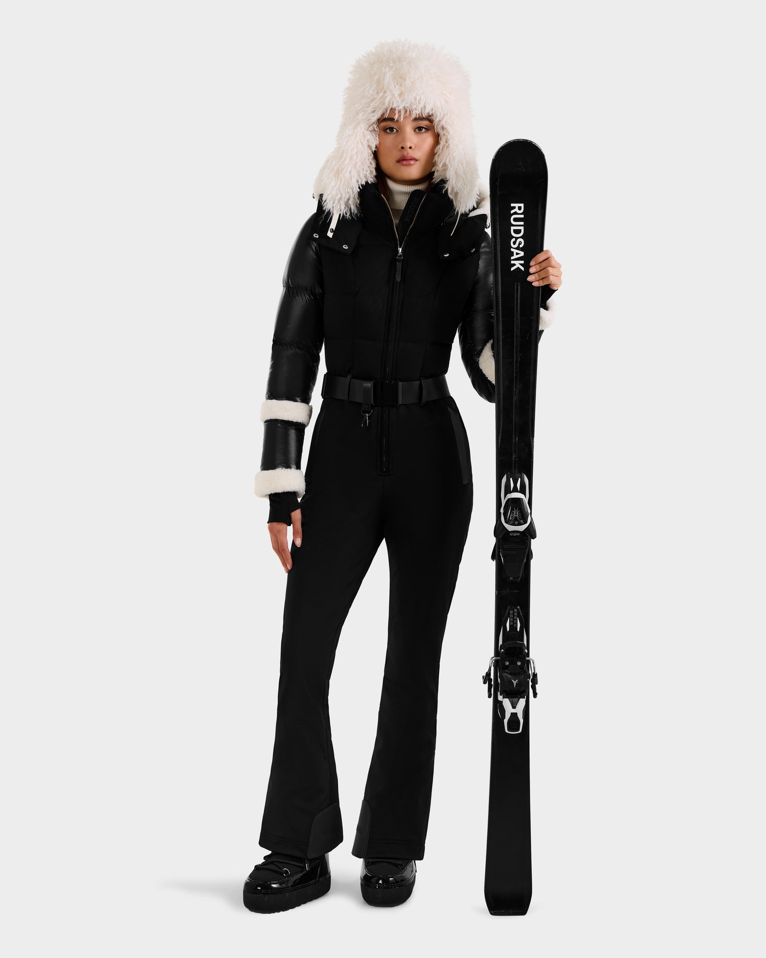 A woman wearing the Rudsak CARMEN - 8125708 Black ski suit, black boots, and a fluffy white hat stands holding black Rudsak skis against a plain white background.