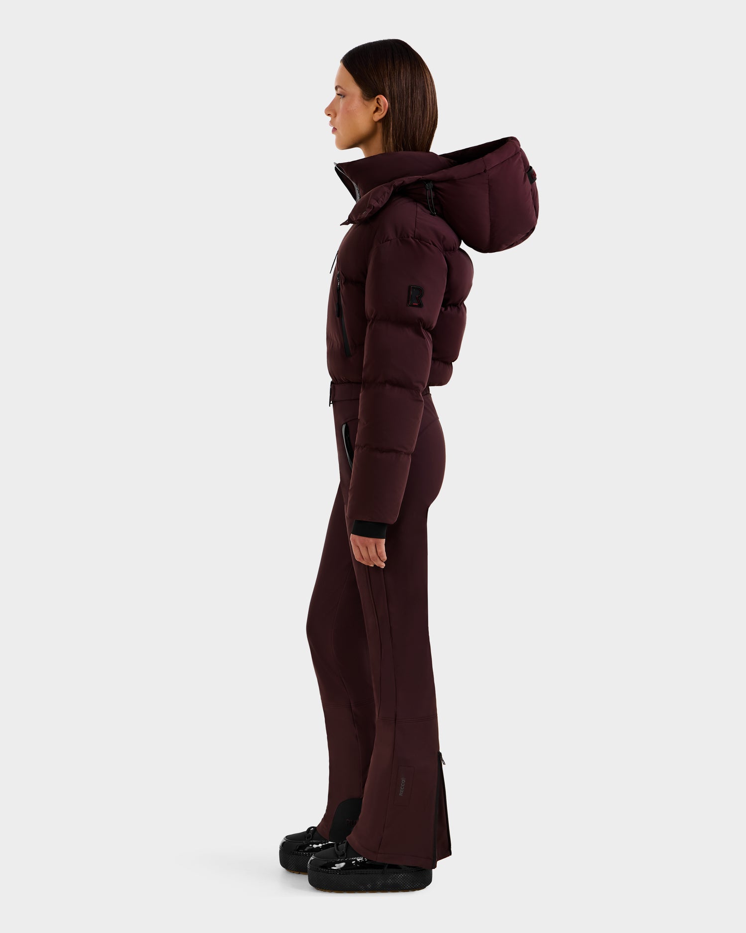 A woman in profile wears the Rudsak DINARA - 8125600 Bordeaux ski suit, featuring a large hood and fitted flared pants. She has straight brown hair, black shoes, and stands against a plain light background.