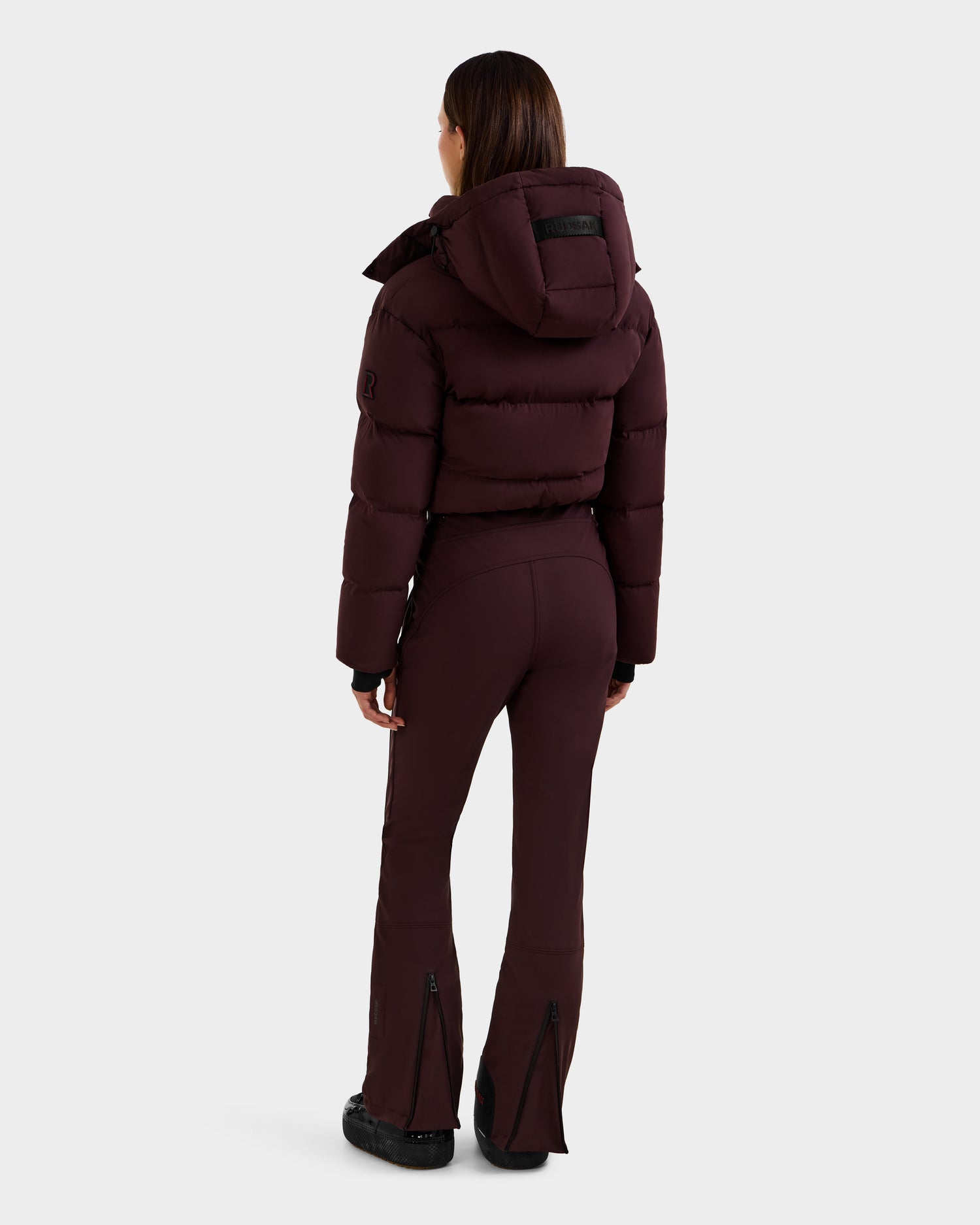 A woman with straight brown hair faces away, wearing the Rudsak DINARA - 8125600 Bordeaux women's ski suit with a hood and fitted pants featuring back ankle zippers, set against a plain light background.
