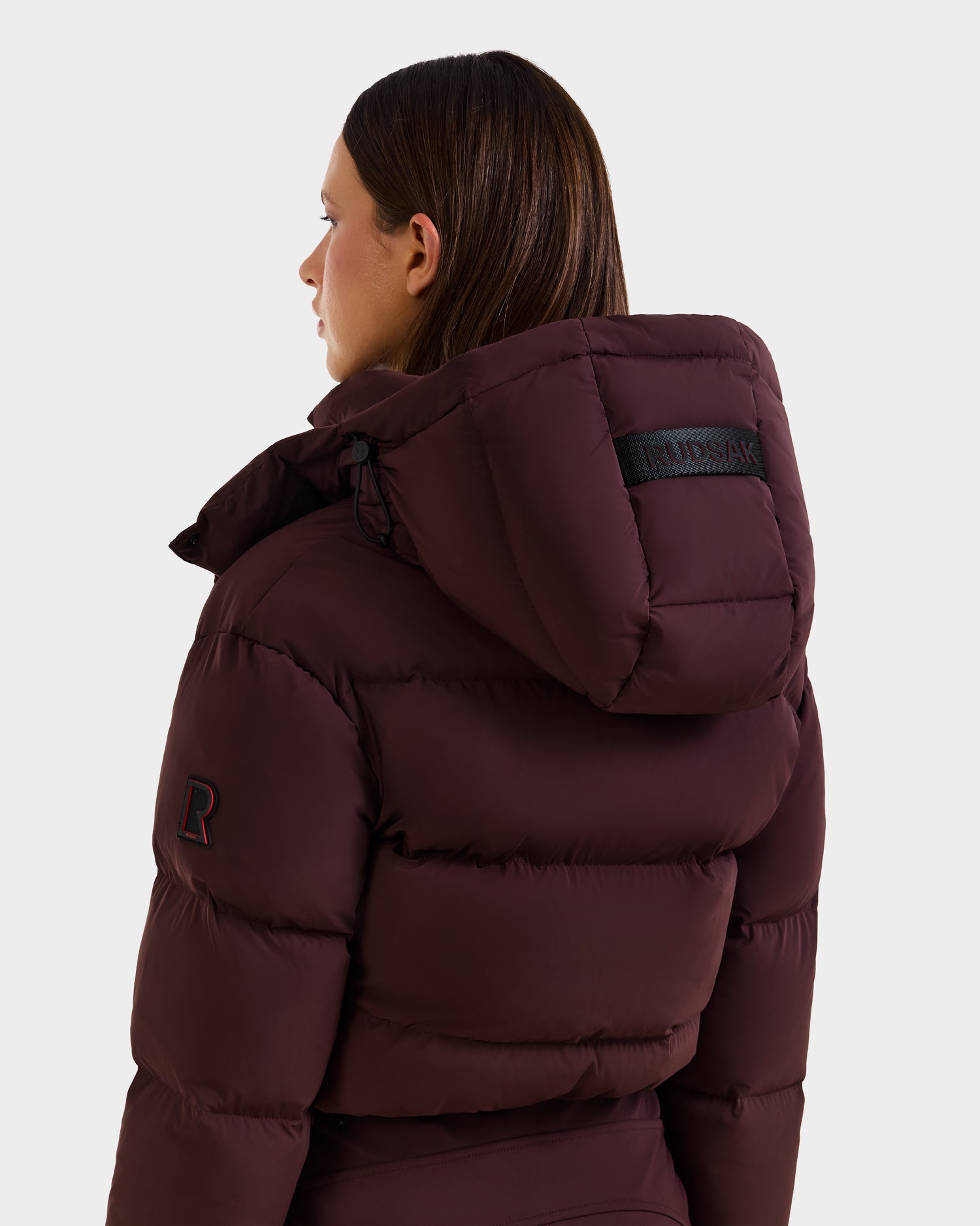 A person is shown from the back wearing the Rudsak DINARA - 8125600 Bordeaux puffer jacket with a hood, black RUDSAK label, and stitched R patch on the sleeve, inspired by a women’s ski suit and set against a white background.
