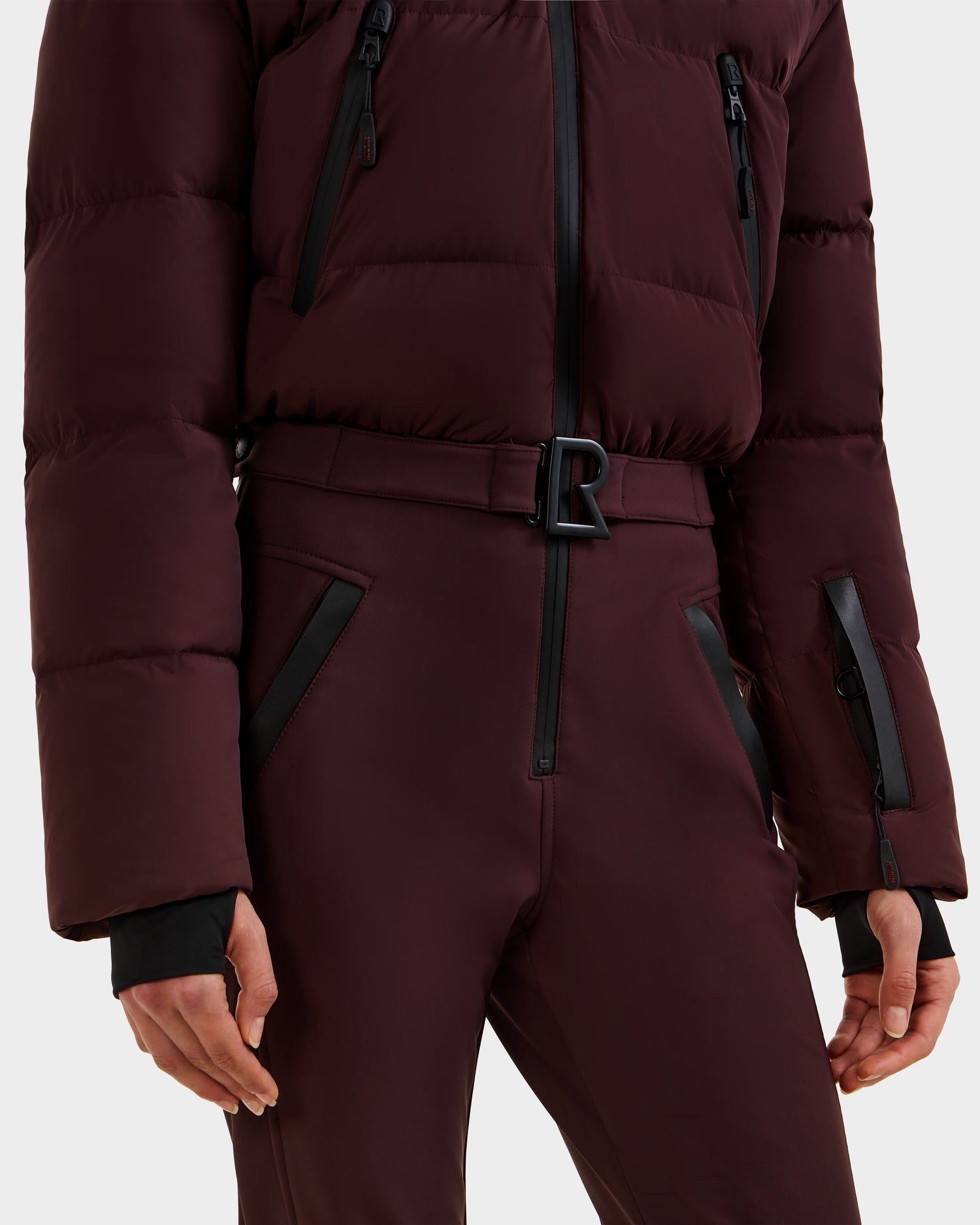 A person wearing the Rudsak DINARA - 8125600 Bordeaux women's ski suit, shown from shoulders to mid-thigh, featuring black zipper details and a belted waist with a black R buckle.