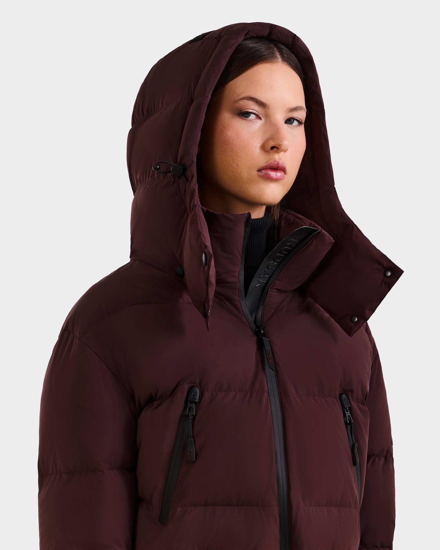A woman wears the Rudsak DINARA - 8125600 Bordeaux puffer jacket with hood up and RECCO rescue technology, looking slightly to the side with a neutral expression against a plain light background.