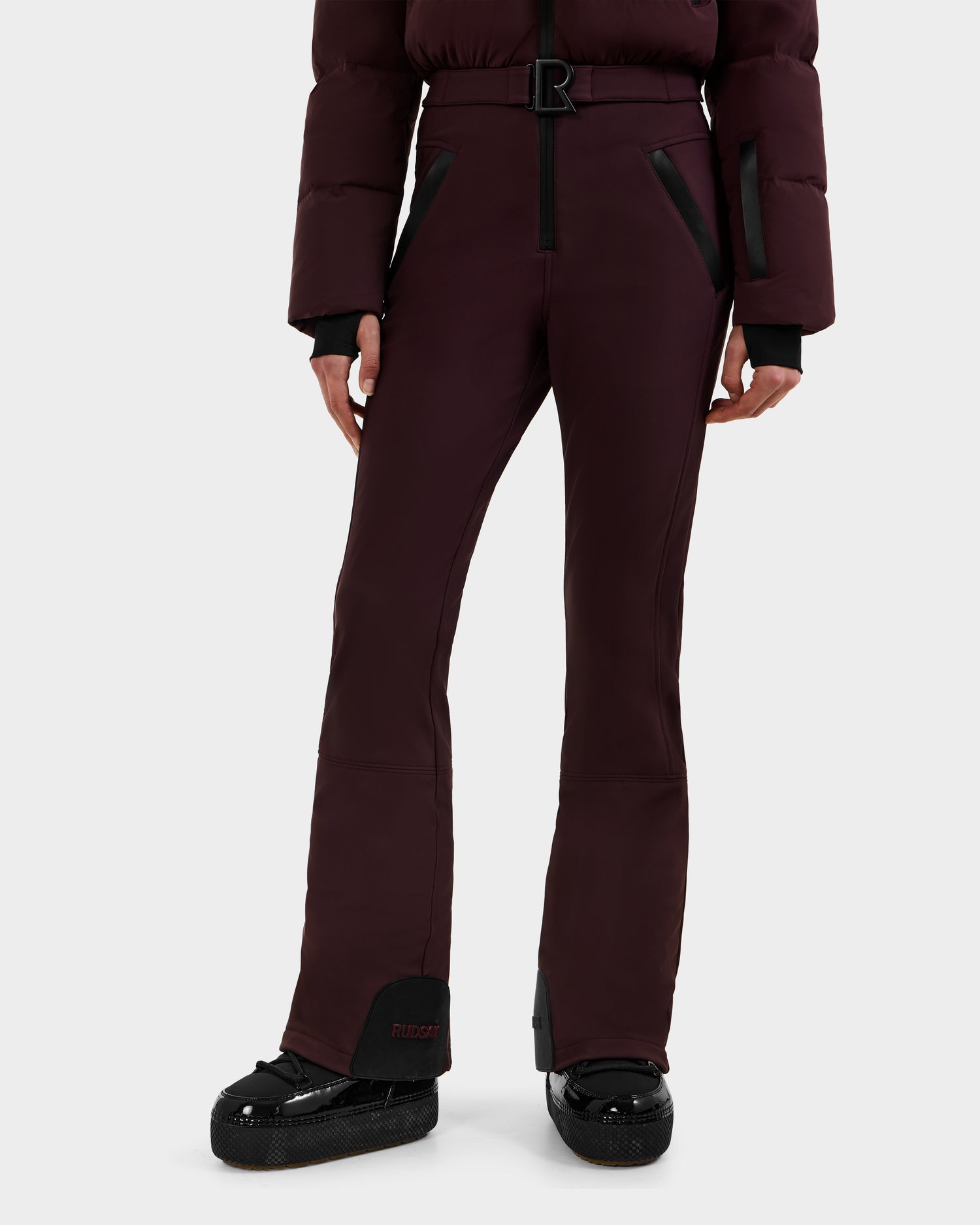 A person wearing Rudsak's DINARA - 8125600 Bordeaux flared pants with black zipper pockets and a matching padded jacket featuring RECCO rescue tech, styled with black platform shoes, stands against a plain white background.