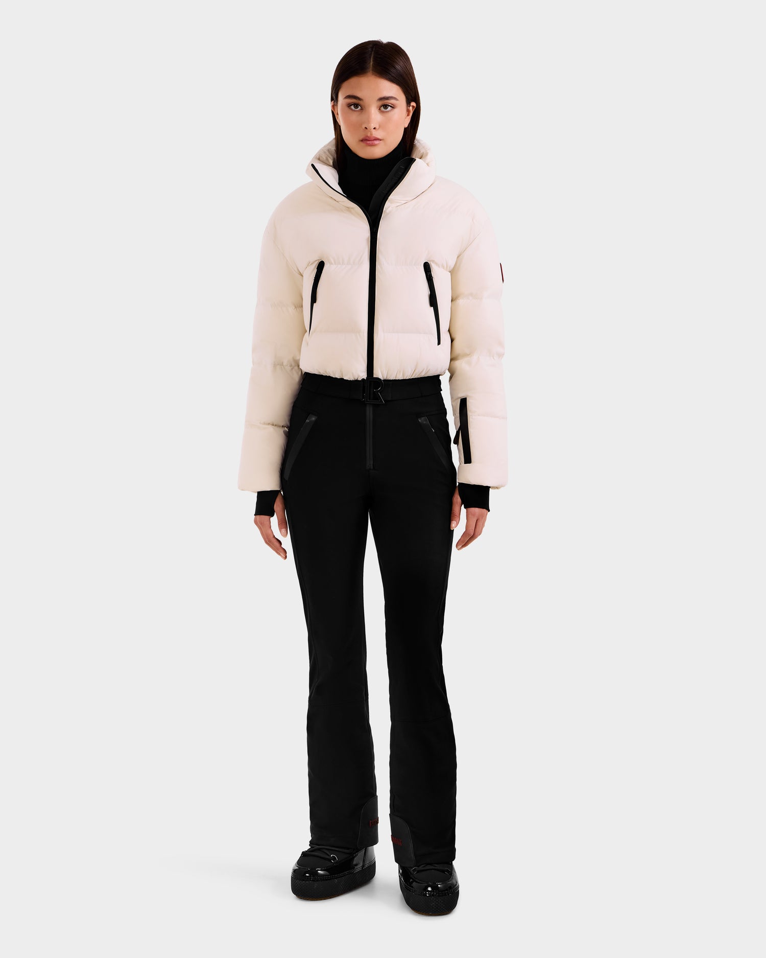 A woman faces forward in the Rudsak DINARA - 8125600 Vanilla white puffer jacket with RECCO rescue tech, black fitted pants, and black shoes against a light gray background. She has straight brown hair and a neutral expression.