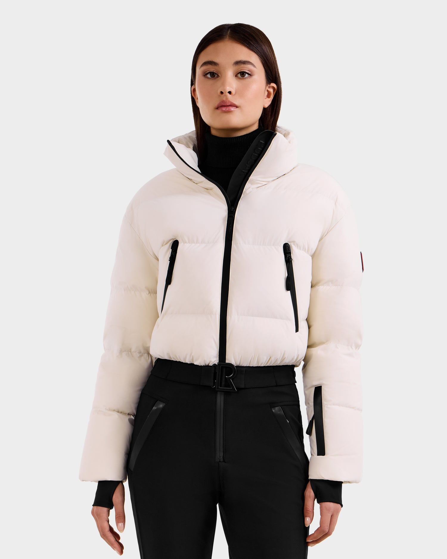 A woman faces forward against a light gray background, showcasing the elegant style of Rudsak's DINARA - 8125600 Vanilla women’s ski suit: a white puffer jacket with black zipper details and high-waisted black pants.