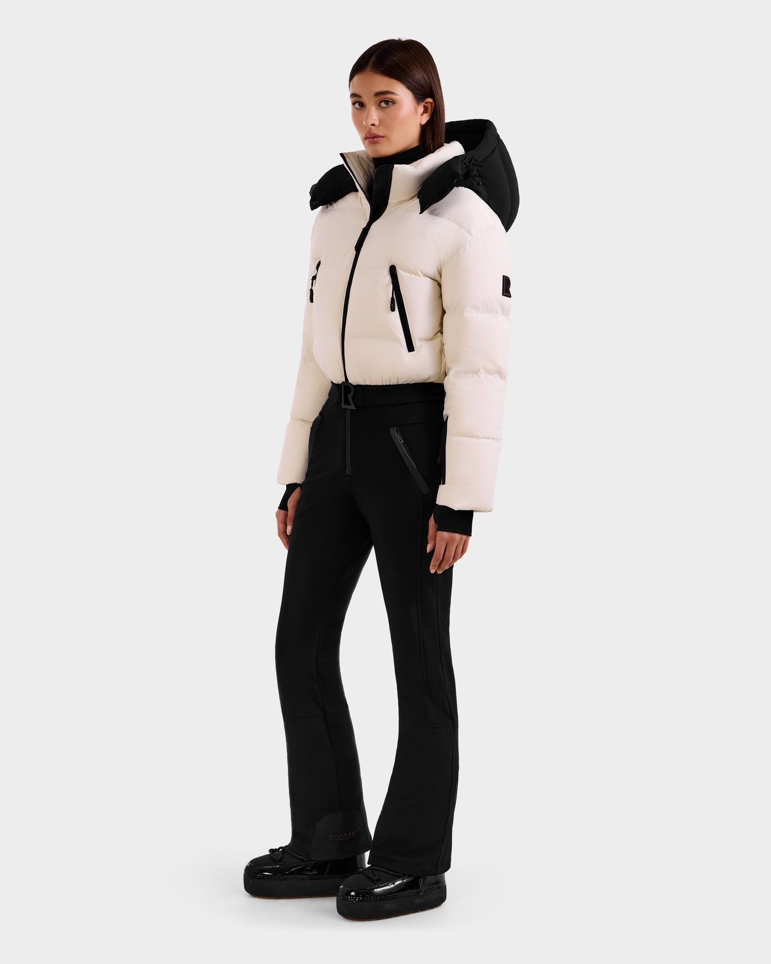 A woman stands slightly to the side against a plain white background, wearing the Rudsak DINARA - 8125600 Vanilla women’s ski suit-inspired white puffer jacket with black accents, paired with black fitted pants and black winter boots.