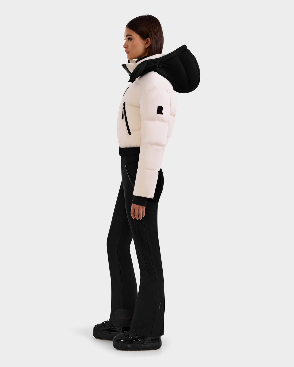 A woman in profile wears the Rudsak DINARA - 8125600 Vanilla white puffer jacket with black accents, paired with black flared pants and RECCO-equipped black shoes, posed against a plain light background.
