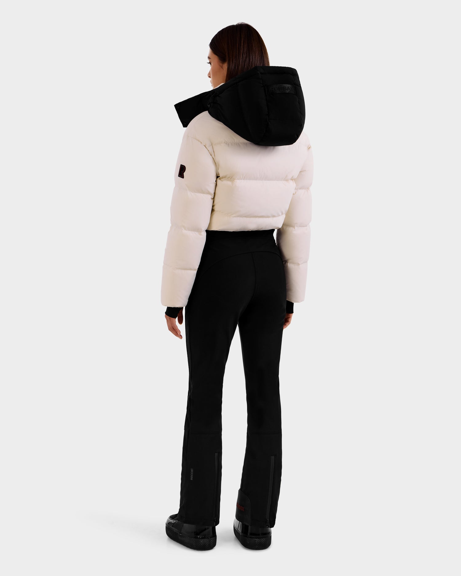 A person stands facing away, wearing the Rudsak DINARA - 8125600 Vanilla white puffer jacket with a black hood, paired with black fitted pants and boots featuring RECCO rescue technology, set against a plain light background.