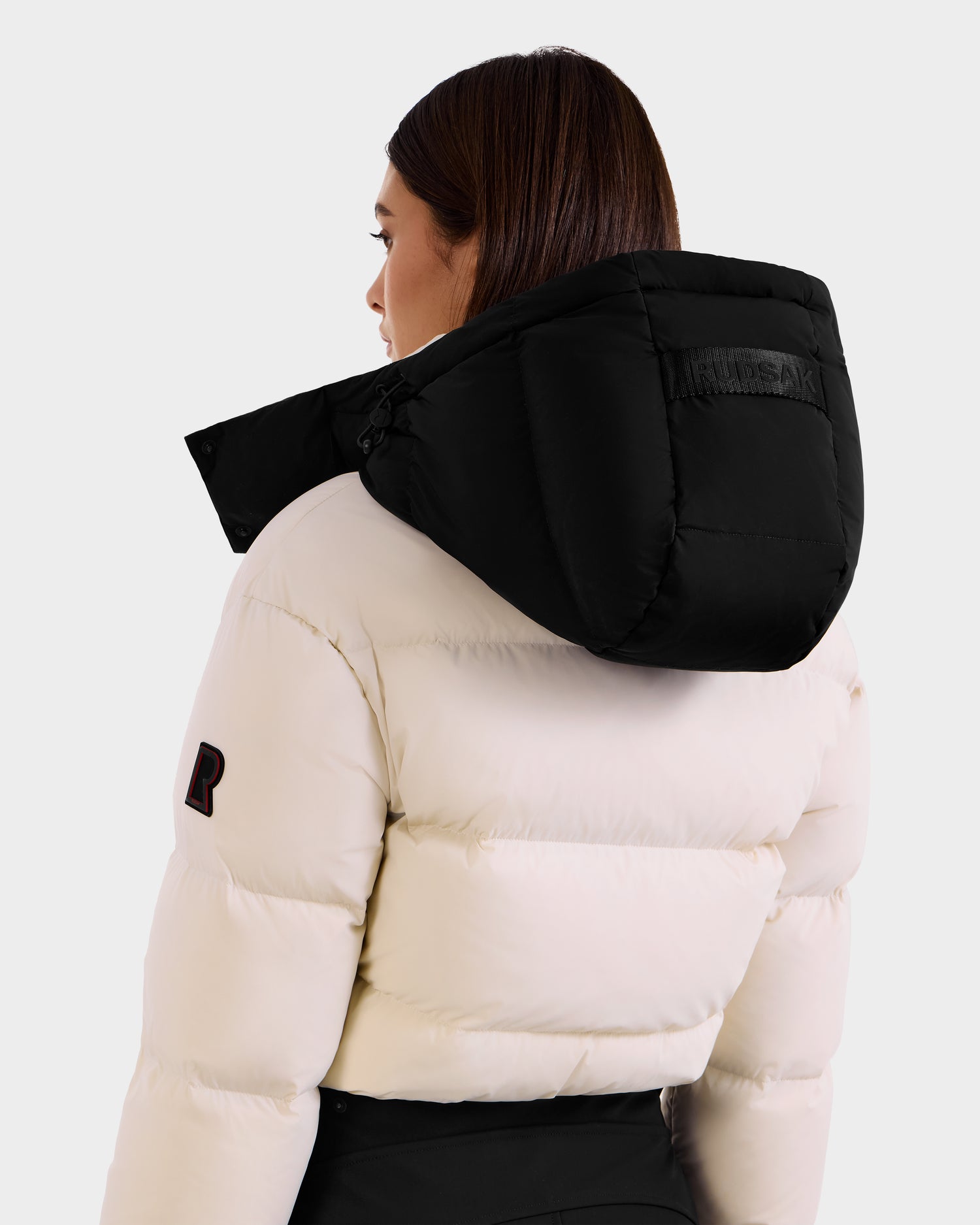 A woman with straight brown hair, seen from the back and in profile against a plain light background, models the Rudsak DINARA - 8125600 Vanilla puffer jacket with a large black hood.