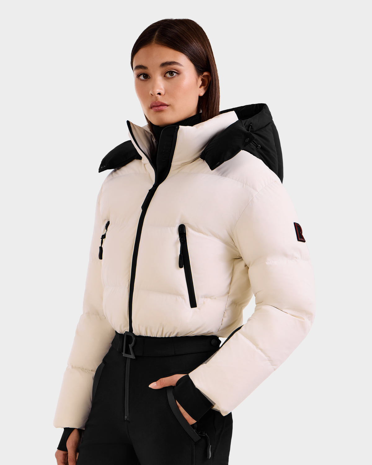 A woman with straight brown hair wears the Rudsak DINARA - 8125600 Vanilla white puffer jacket with RECCO® rescue tech and black zippers, paired with black pants, standing against a plain light background.