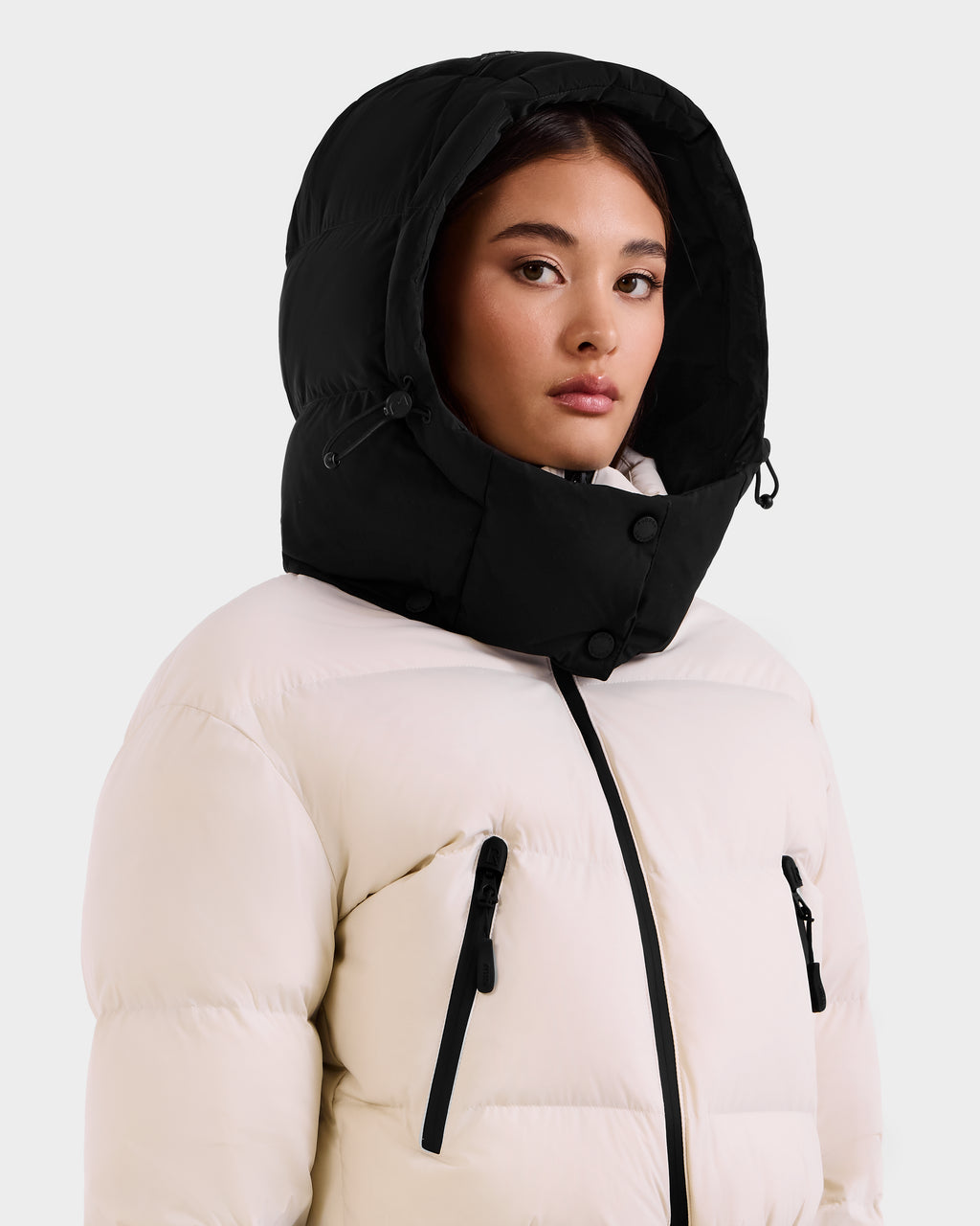 A person wearing the Rudsak DINARA - 8125600 Vanilla puffer jacket with a black hood, black zippers, and buttons looks to the side against a plain light background. The jacket features RECCO rescue technology for added safety.