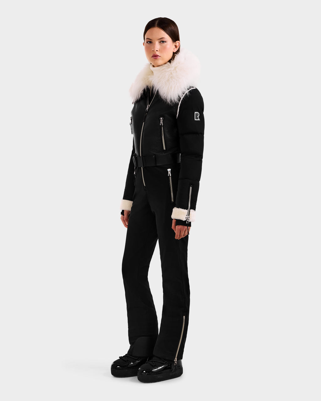 A woman models the Rudsak CHYNTIA - 8125611 Black ski suit, featuring a large white fur collar, zippered accents, and matching black snow boots. A plain light backdrop highlights this chic, high-fashion outfit.