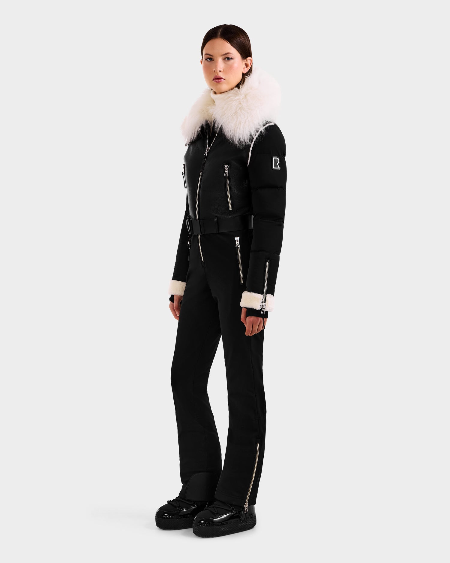A woman models the Rudsak CHYNTIA - 8125611 Black ski suit, featuring a large white fur collar, zippered accents, and matching black snow boots. A plain light backdrop highlights this chic, high-fashion outfit.